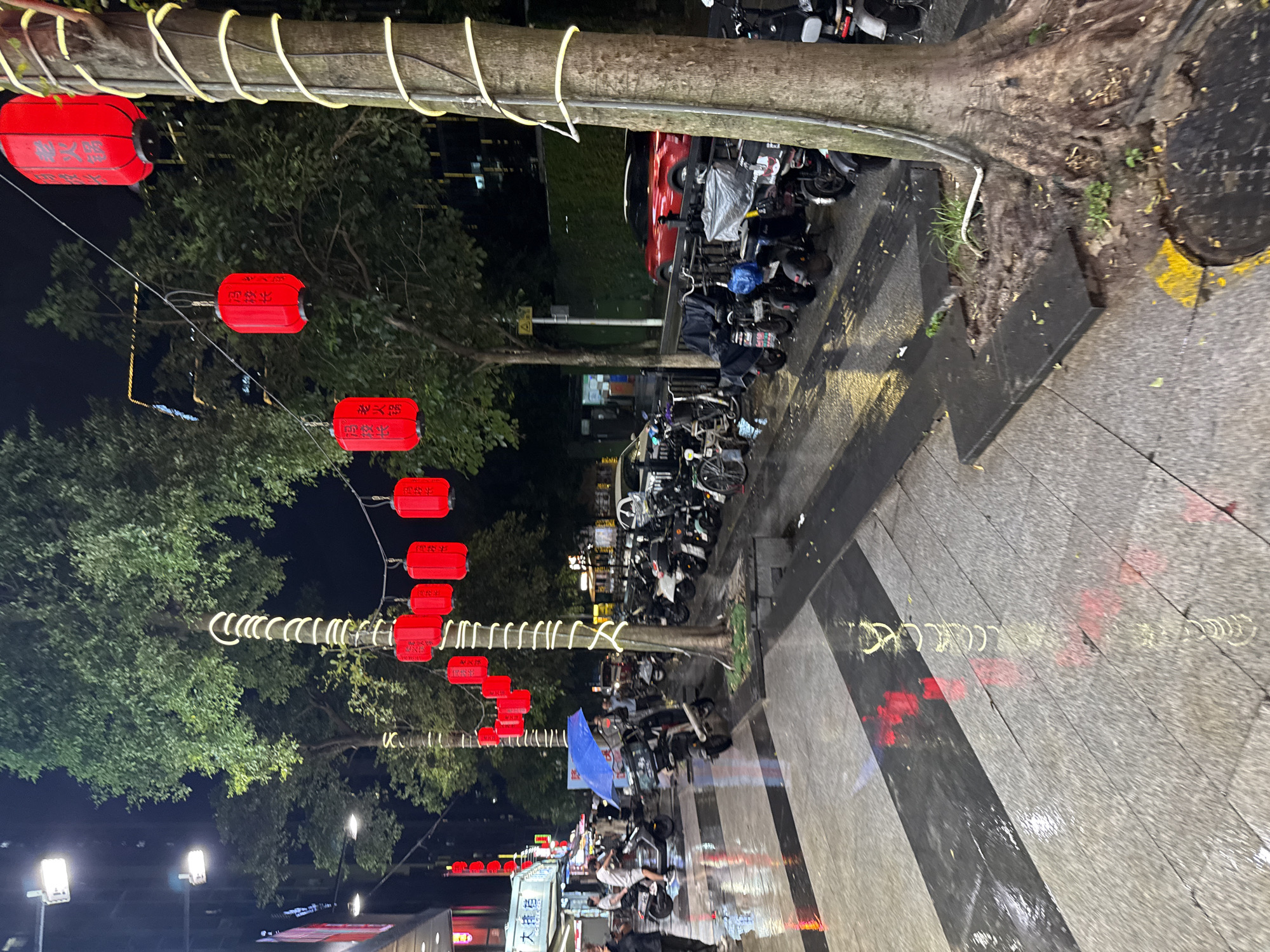 The image depicts a nighttime scene on a wet city street in Haizhu District, China, likely after rainfall, as evidenced by the reflections on the pavement. Red lanterns are strung across the street and suspended from trees, some of which are illuminated with yellow spiral lights. A variety of parked electric scooters and motorcycles line the side of the street. Several individuals are visible, including a person holding a blue umbrella, and others appearing to walk or stand near the scooters. Buildings with illuminated signage are present in the background, and streetlights provide additional illumination. The ground is paved with stone tiles and asphalt, with some areas appearing darker due to wetness and light reflections.