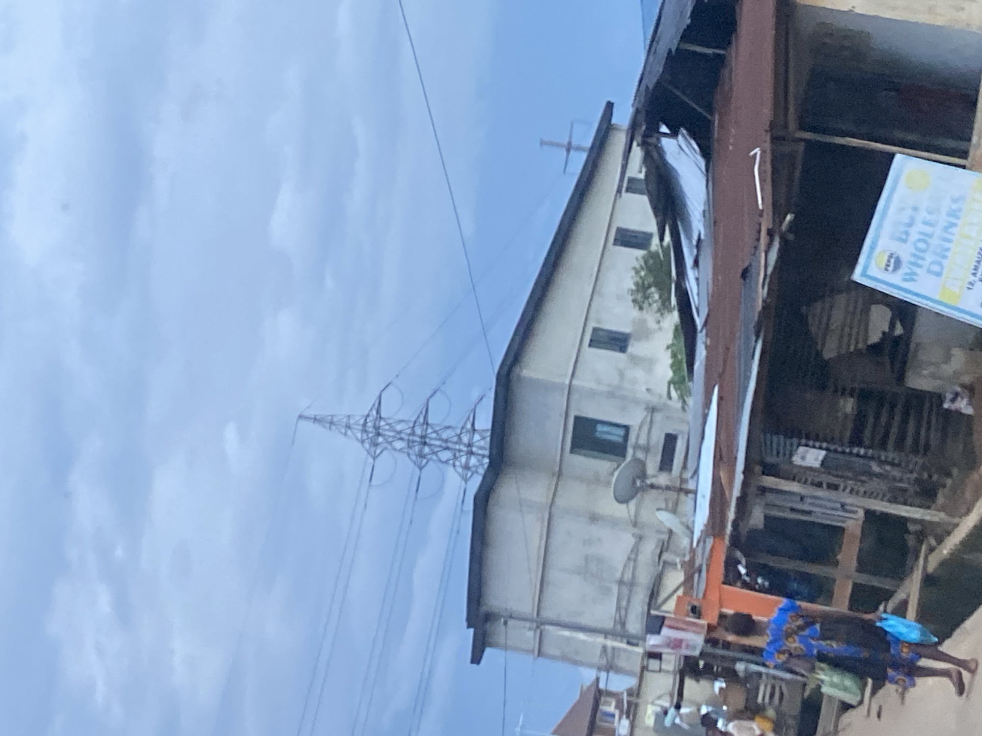 The image depicts a street scene in Ikorodu, Nigeria. A woman, seen from behind, walks on a dirt path towards the right of the frame. She is wearing a blue and black patterned dress and carrying two shopping bags, one green and one blue. In the background, a multi-story building with visible windows stands prominently. Adjacent to it, a structure with a corrugated metal roof appears to be a market stall or shop. Overhead, power lines and a large transmission tower dominate the sky, which is filled with overcast clouds. The overall impression is of an urban environment with elements of daily life and infrastructure.