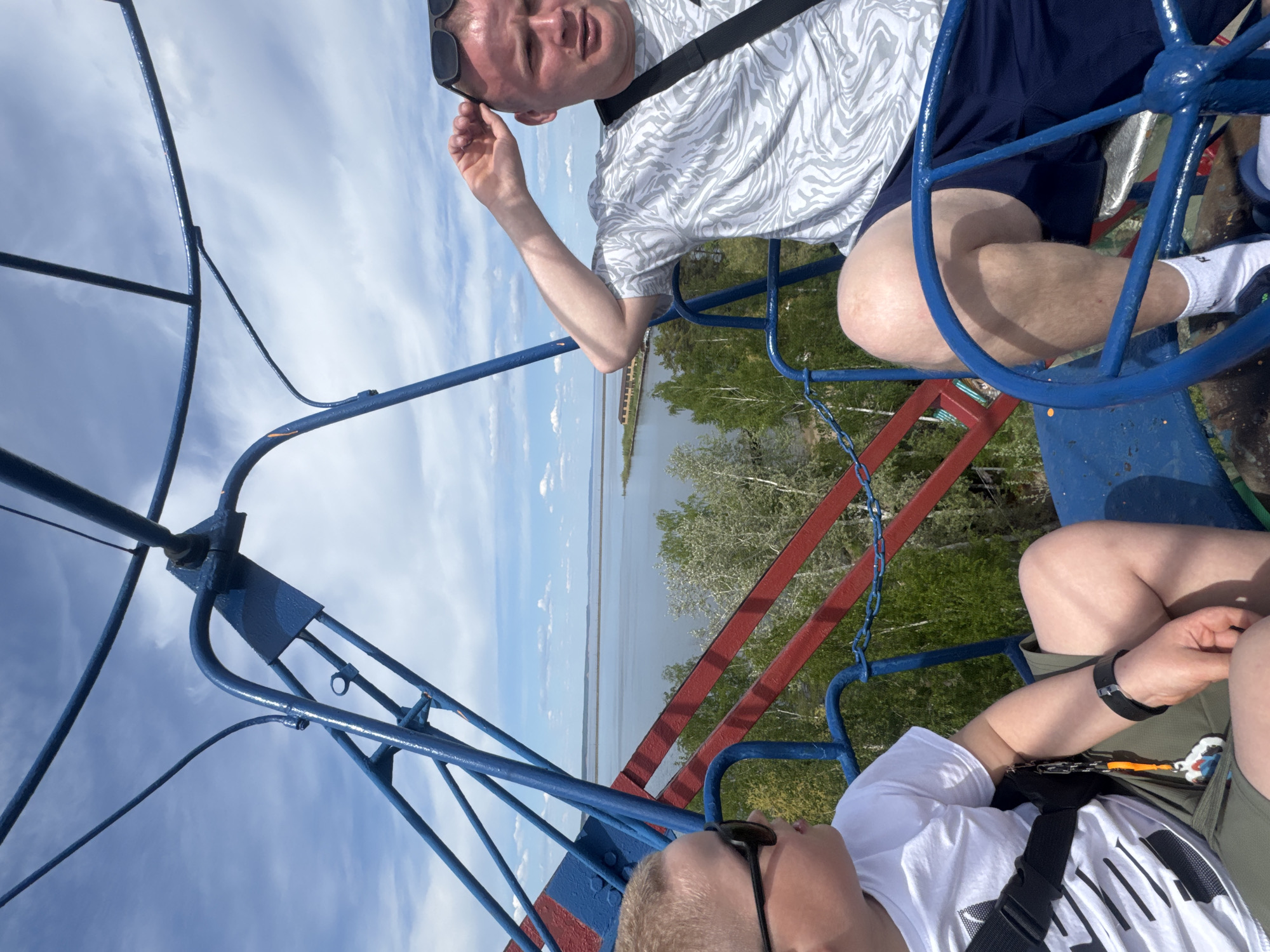 Two men are sitting on a blue and red Ferris wheel at the beach in the city of Chaikovsky, Russia. The Ferris wheel seats are blue, with red supports. One man is wearing a white and gray patterned shirt, dark shorts, a black shoulder strap, and white socks. He has sunglasses propped up on his head and is holding his hand up. The other man is wearing a white t-shirt, khaki shorts, and a black shoulder strap, with sunglasses on his face. In the background, a body of water is visible, with a shoreline and trees. The sky is cloudy.