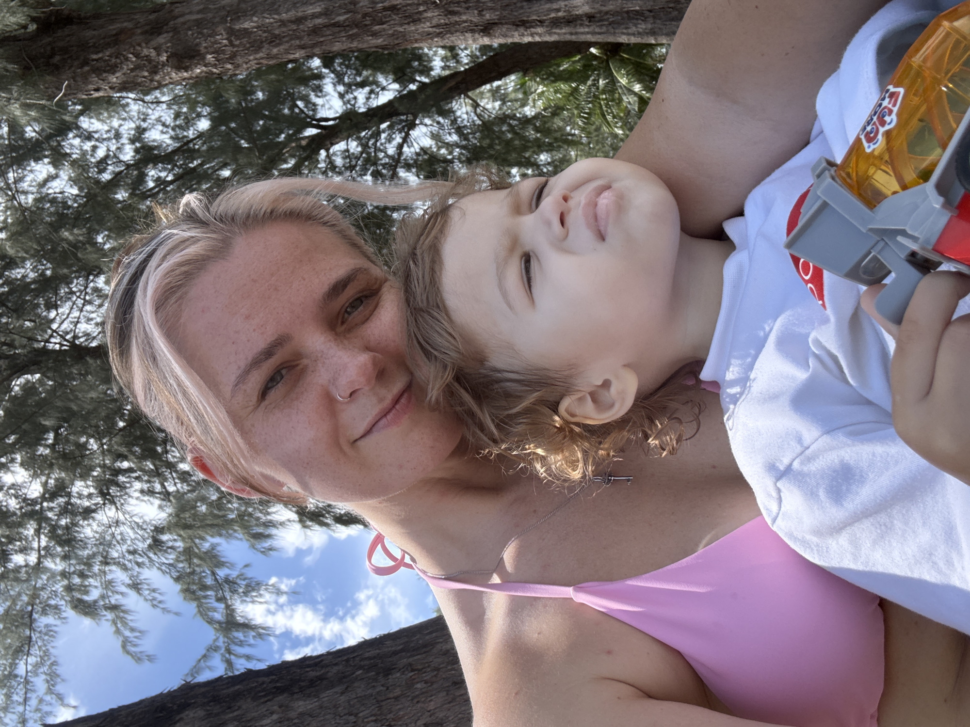 In Choeng Thale, Thailand, a woman with blonde hair and freckles, wearing a pink bikini, is holding a young child. The child, with curly brown hair, is looking away from the camera. They are seated outdoors, with trees and a bright blue sky with white clouds visible in the background. The child is holding a toy.