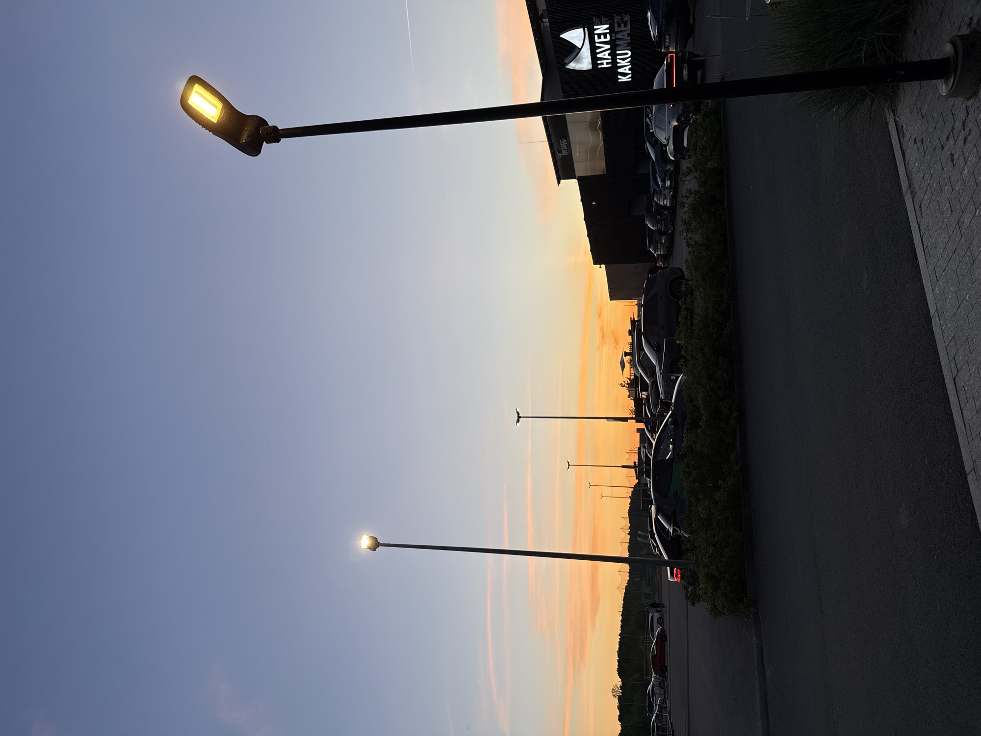 The image depicts a parking lot at dusk with a warm, orange and pink sunset visible in the background. Several cars are parked in the lot, and there are multiple lampposts, one of which is illuminated. In the background, a modern building with illuminated signage that reads "HAVEN KAKUMÄE" is visible. The scene is located in Tallinn, Estonia. There are no people clearly visible in the image. The overall atmosphere is calm and serene.