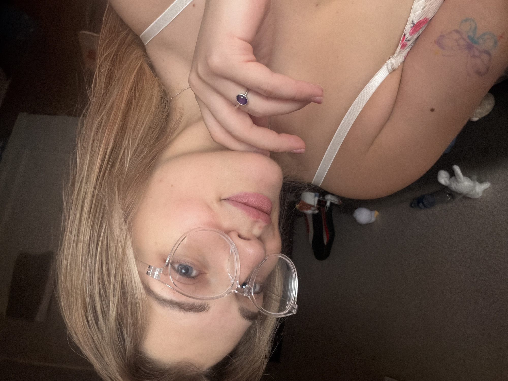 A close-up, angled shot shows a person with blonde hair and glasses. The person is wearing what appears to be a light-colored bra or top with thin straps. A ring with a purple stone is visible on one of the person's fingers. A tattoo resembling a flower or butterfly is on the person's shoulder/upper arm. The background is blurry and indistinct but appears to be a carpeted floor. There is not any evidence of the city of Naberezhnye Chelny, Russia.