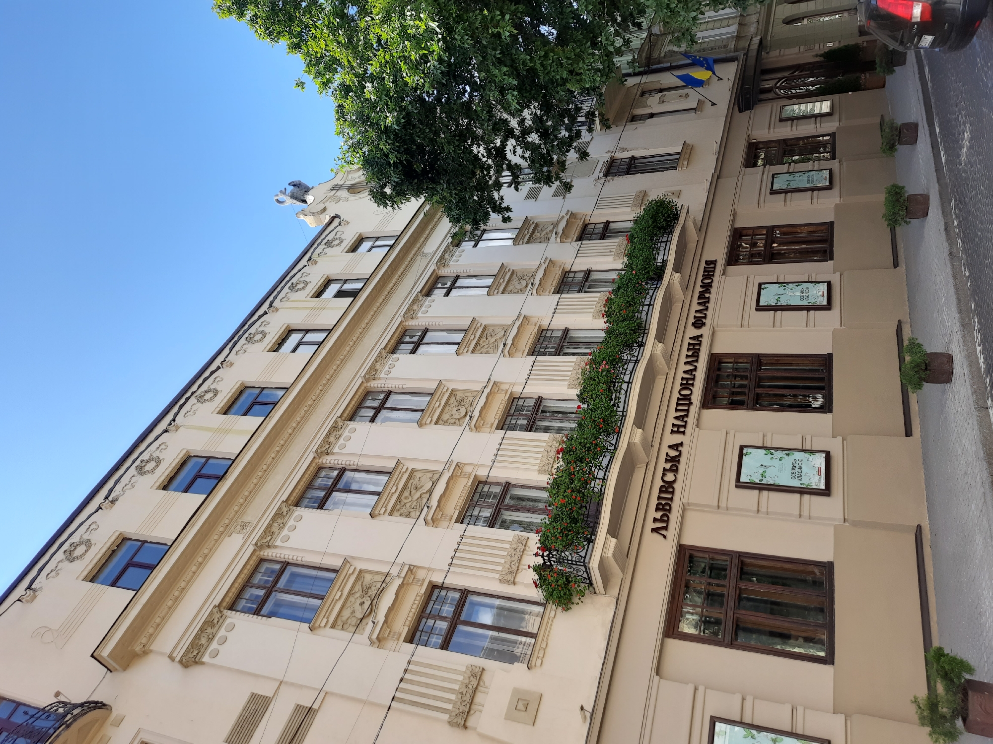 The image captures the facade of a grand, multi-story building in Lviv, Ukraine, identified by the Cyrillic inscription "ЛЬВІВСЬКА НАЦІОНАЛЬНА ФІЛАРМОНІЯ" (Lviv National Philharmonic). The building is painted in light beige and cream tones, with ornate architectural details, including decorative carvings above the windows and a balcony overflowing with red flowers. The windows are dark brown, and the roofline features intricate friezes. A prominent sculptural element, possibly a bird or mythological creature, crowns the building's peak. Two flags, the Ukrainian flag and the European Union flag, are displayed on the right side of the building. The lower part of the building features glass-enclosed display cases with posters. Potted plants line the cobblestone sidewalk in front of the building, and a dark-colored car is partially visible on the right. The sky is a clear, bright blue, and a large, leafy tree stands to the right, partially obscuring the building. The overall impression is one of a well-maintained historic cultural institution in an urban setting.