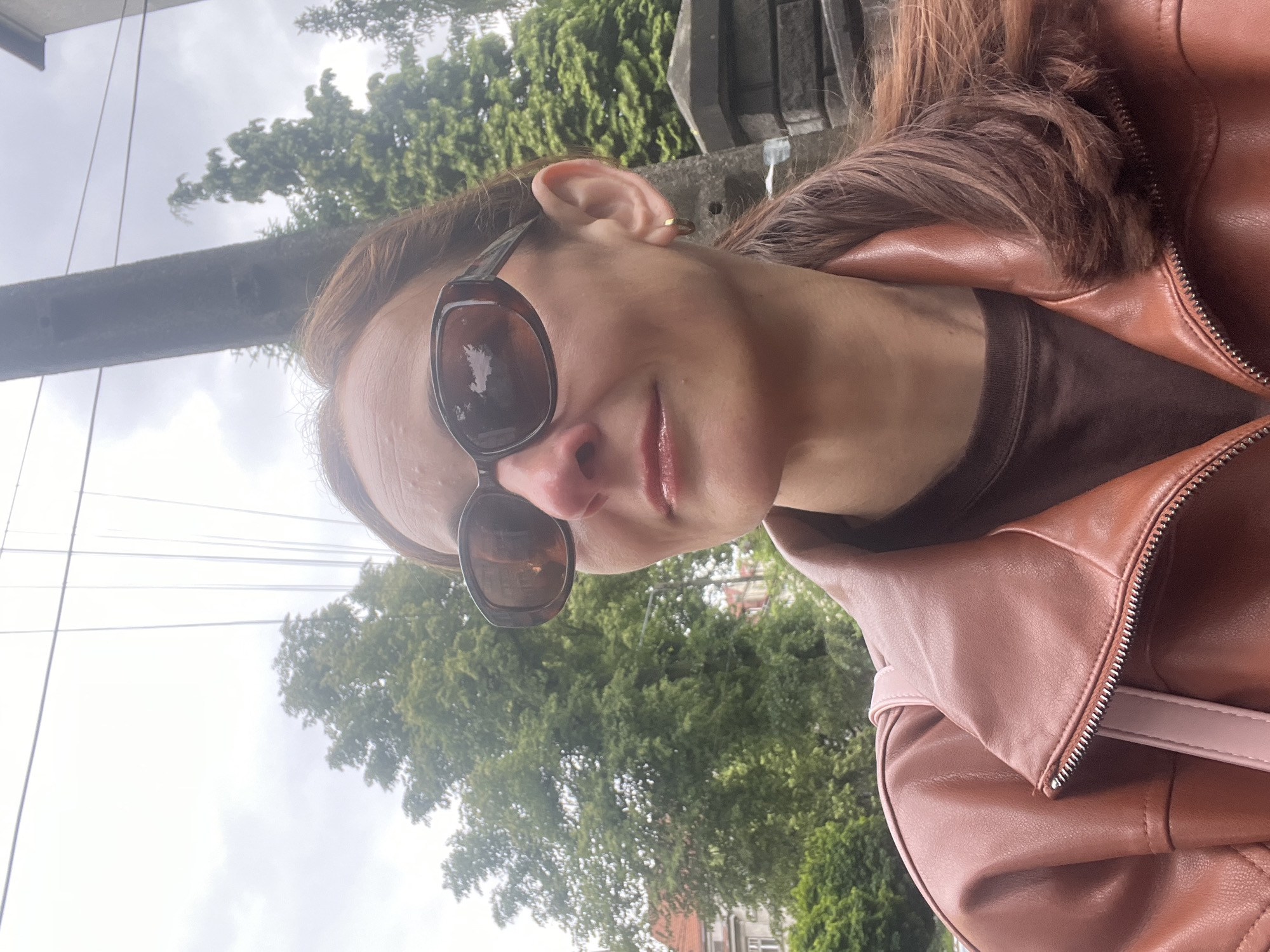 A woman with auburn hair pulled into a ponytail is taking a selfie outdoors. She is wearing large, dark sunglasses, a brown t-shirt, and a reddish-brown leather jacket. A pink strap from a bag is visible over her shoulder. She has a small gold hoop earring in her right ear. Behind her, there are trees, a utility pole with wires, and part of a stone wall. The sky is overcast.