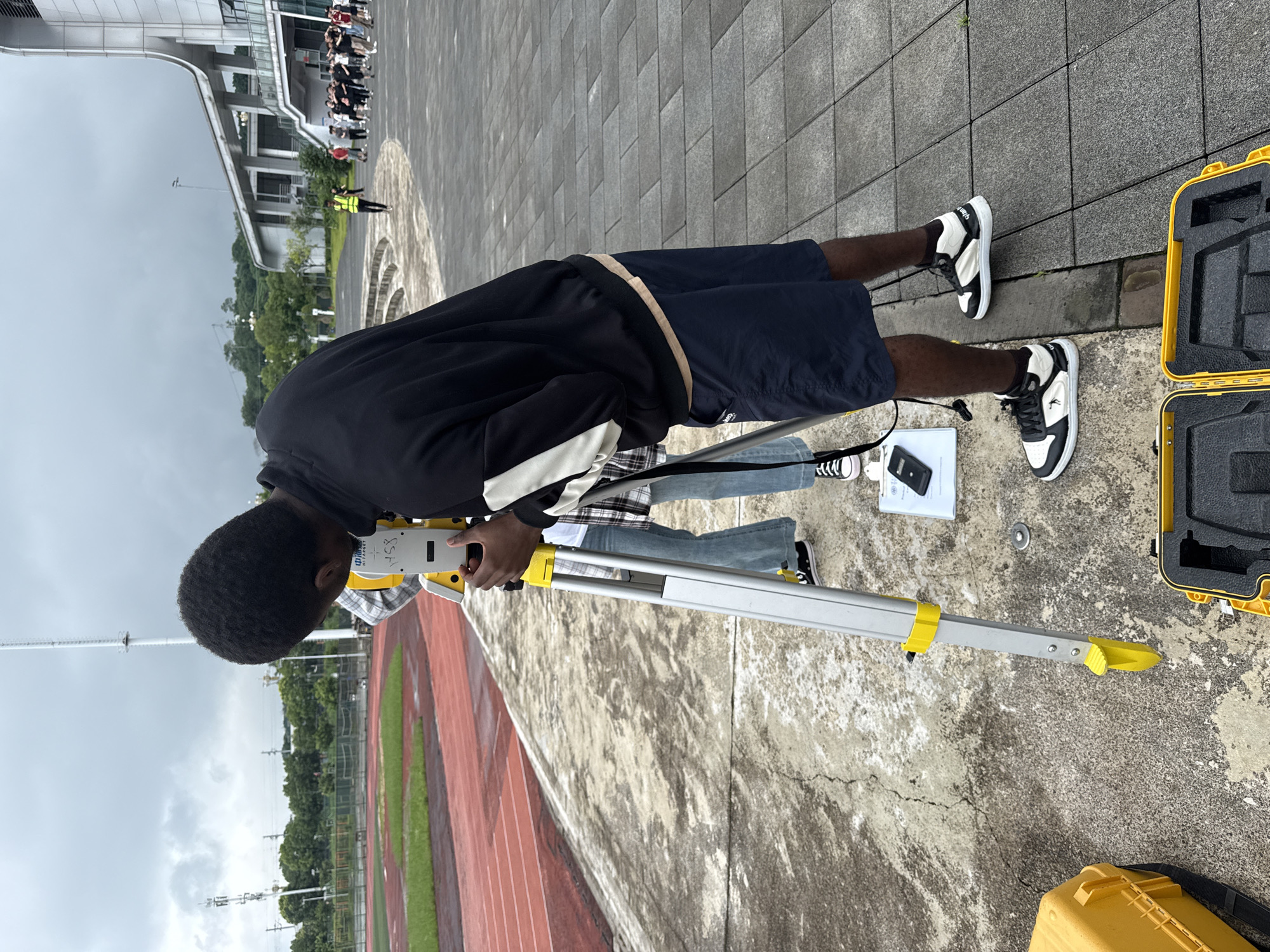In Jiangjin, China, a young Black man is operating a surveying instrument, a total station, on a paved area next to a running track. He is wearing a black zip-up jacket with white stripes on the sleeves, navy blue shorts, and black and white sneakers. Behind him, another person, partially visible, is wearing jeans and a plaid shirt, also looking at the surveying equipment. The instrument is mounted on a tripod, and a yellow carrying case is open nearby. In the background, a stadium with tiered seating and a large modern building are visible under a cloudy sky. Other people are gathered in the distance near the building. The environment appears to be an outdoor sports facility or campus area.
