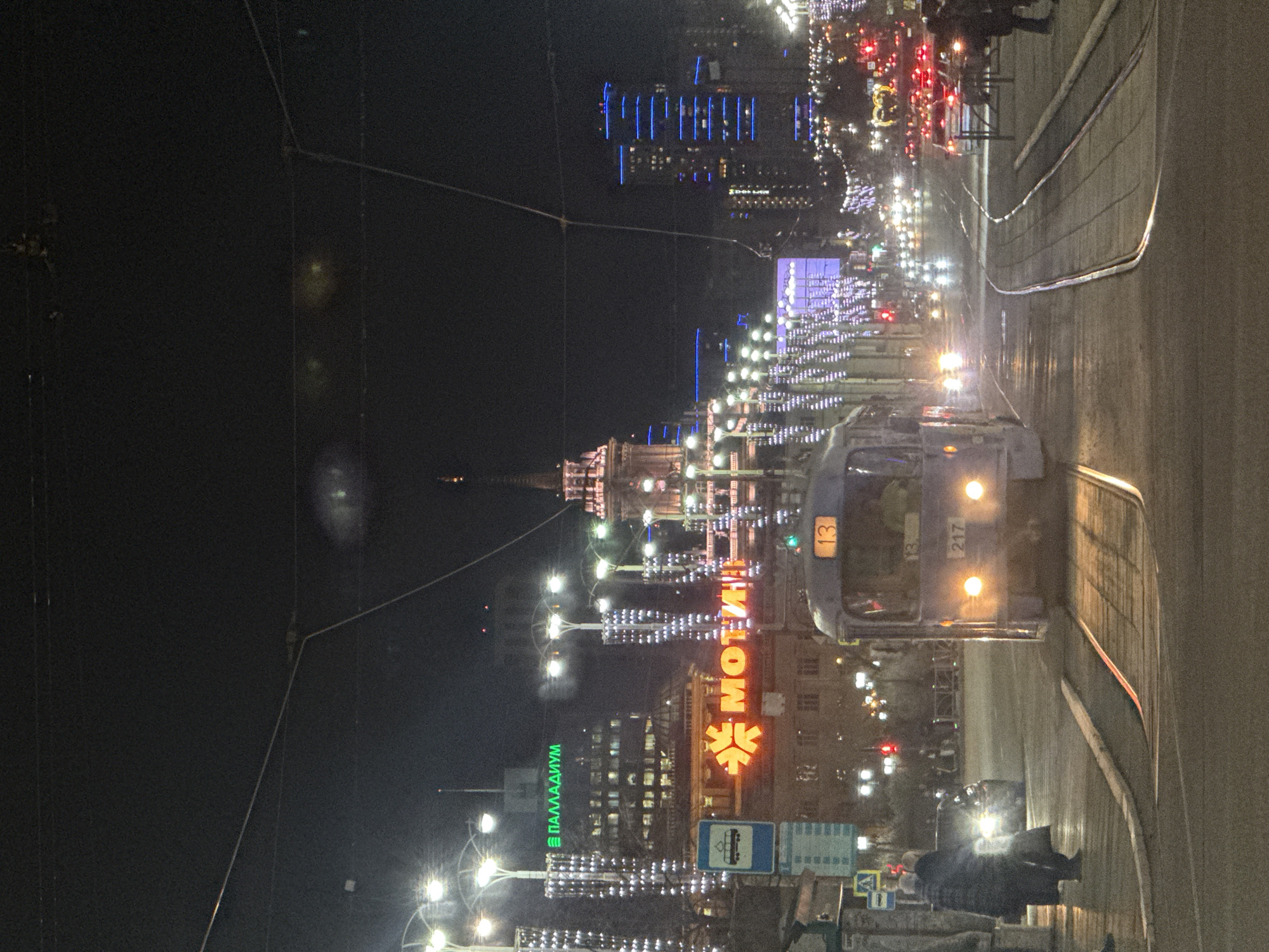 The provided image, captured at night in Yekaterinburg, Russia, shows a bustling city street with a tram as a central subject. The original image is rotated 90 degrees clockwise.
In the upright view, a tram, numbered "13" on its front and "217" on its side, occupies the right side of the frame, appearing to be stopped or moving slowly along its tracks. Its headlights are on, reflecting off the wet road surface. The street extends into the distance, filled with traffic, evidenced by blurred red and white vehicle lights.
The scene is brightly illuminated by various artificial lights against the dark night sky. Streetlights and several buildings are decorated with numerous white, possibly festive, string lights. In the mid-ground, a grand, older building with a spired roof is prominently lit, showcasing its architectural details. Further back, modern high-rise buildings display illuminated windows and some feature distinct blue accent lighting.
Several visible signs identify businesses or points of interest. A large, orange neon sign clearly displays "МОТИВ" (MOTIV) alongside a unique star or snowflake-shaped logo. Below and slightly to the left, another building bears a green neon sign spelling "ПАЛЛАДИУМ" (PALLADIUM). A standard blue bus stop sign with a white bus icon is visible on the sidewalk. Overhead, a complex network of wires for the tram system is silhouetted. Several indistinct figures of people can be seen on the sidewalk, adding to the urban activity. The overall impression is one of a vibrant, illuminated city after dark.