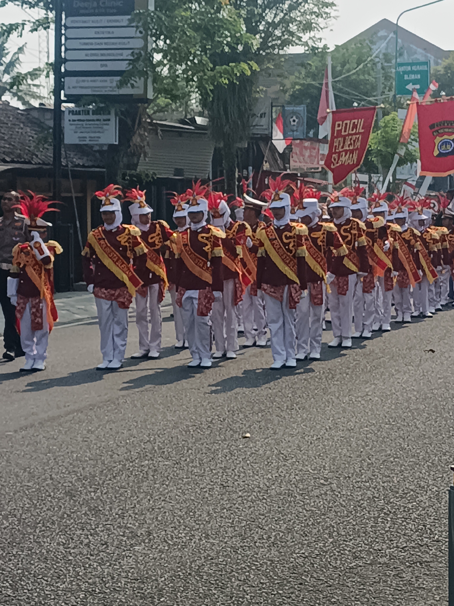 The image shows a marching band in uniform, walking on a paved street in Sleman, Indonesia. The band members are wearing maroon jackets with gold trim, white pants, and white helmets with red plumes. They are also carrying instruments, such as drums and bugles. In the background, there are buildings, trees, and street signs. One sign reads "Polcil Polresta Sleman," indicating the location. The weather appears to be sunny.