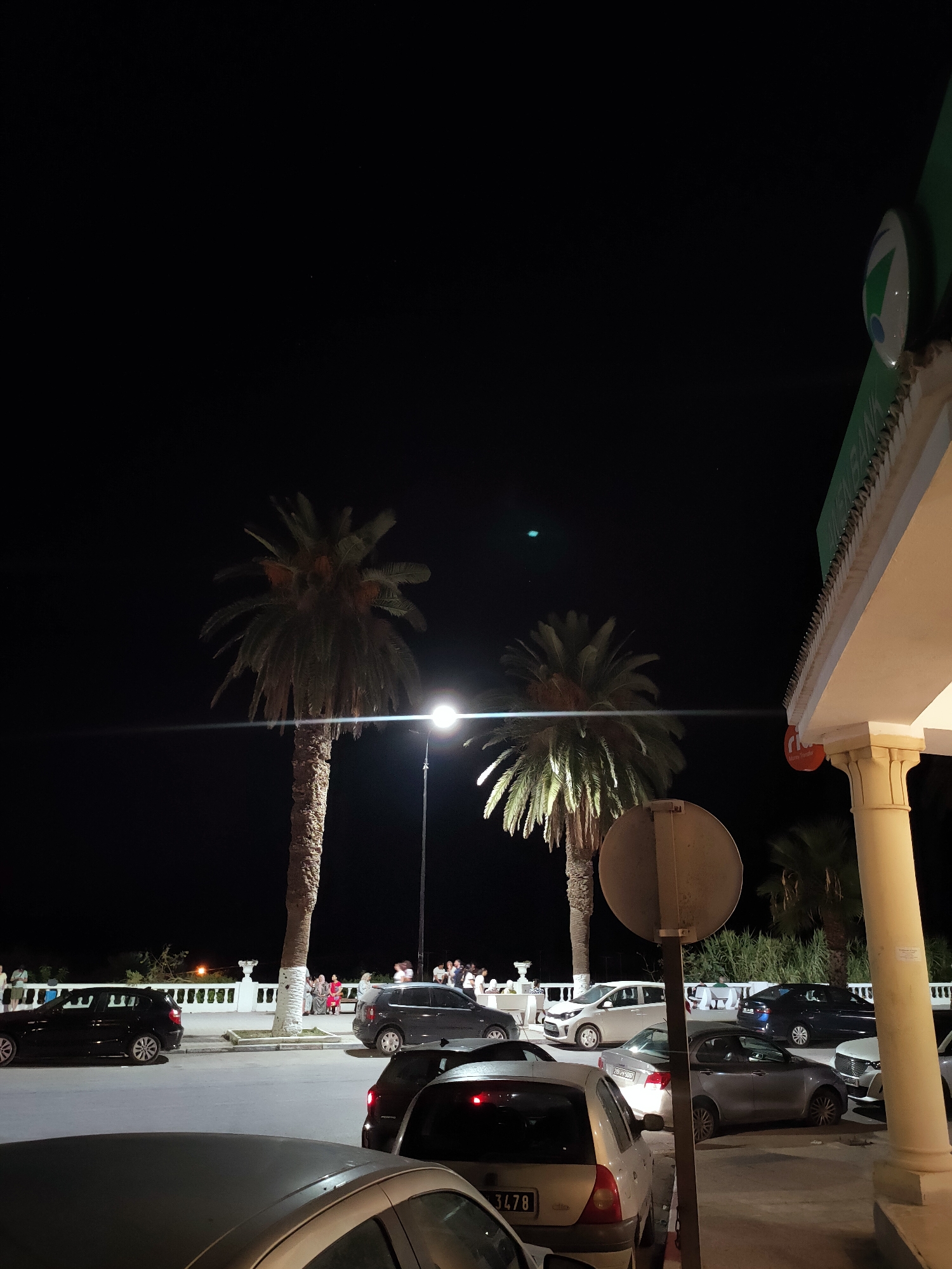 In the city of Tunis, Tunisia, at night, the image captures a street scene. Two palm trees stand prominently, illuminated by a nearby street lamp. Several cars are parked on the street and a sidewalk area, including a dark SUV, a grey sedan, and a white car. In the background, a white railing separates the sidewalk from a slightly elevated area where a group of people are gathered, some sitting and others standing. The facade of a building with a green awning and a distinctive circular logo is visible on the right side of the frame. The sky is dark, indicating nighttime.