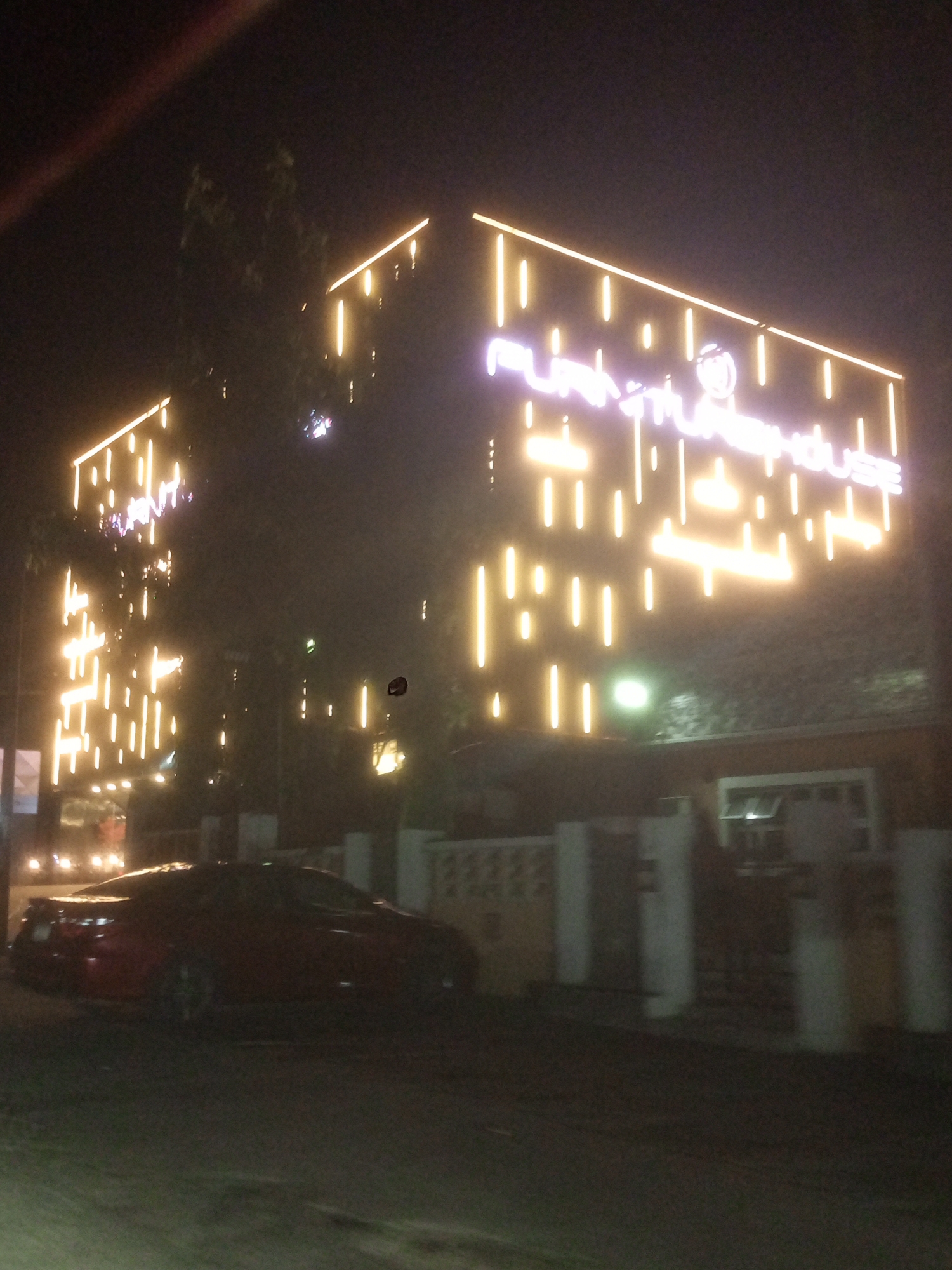 The image shows a building illuminated with decorative vertical LED lights in Gwarinpa, Nigeria. The building has a sign reading "aura warehouse" in all lowercase letters. A red sports car is parked in front of the building. A fence runs along the perimeter of the property. The scene appears to be at night, and the overall lighting is poor. Trees are visible in the background. The road in the foreground is dark. The image appears to be taken from a low angle.