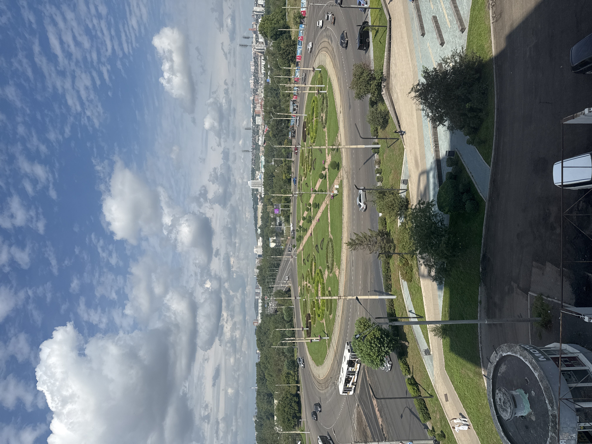 An elevated, wide-angle shot captures a circular intersection in Krasnoyarsk, Russia, on a partly cloudy day. The intersection is a prominent feature with a large, manicured green roundabout at its center, adorned with patterned shrubbery and small trees. Several cars and a white bus are visible navigating the asphalt roads surrounding the roundabout. In the distance, a cityscape unfolds with numerous buildings and infrastructure, including roads and bridges, set against a backdrop of lush green trees and rolling hills. A modern plaza with tiered seating and decorative elements is situated to the right of the roundabout. In the foreground, a round, grey structure with the partial word "MOTO" visible on its side is present, with a dark asphalt area surrounding it. Two individuals are walking on a grassy area to the left of the plaza.