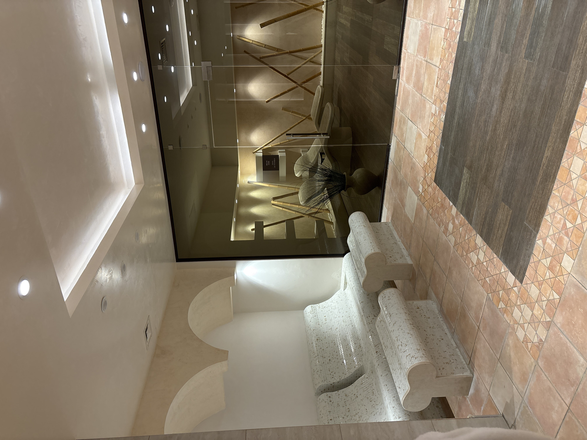 The image shows a spa relaxation area with mosaic-tiled benches and a room with lounge chairs visible through a glass wall. The room is decorated with bamboo stalks and has a rustic aesthetic. The flooring is a combination of terracotta tiles and dark wood planks. The lighting is soft and ambient, with recessed ceiling lights and accent lighting. The scene is clean and tranquil, suggesting a place for rest and rejuvenation. The location is identified as a spa in the city of Razliv, Bulgaria.