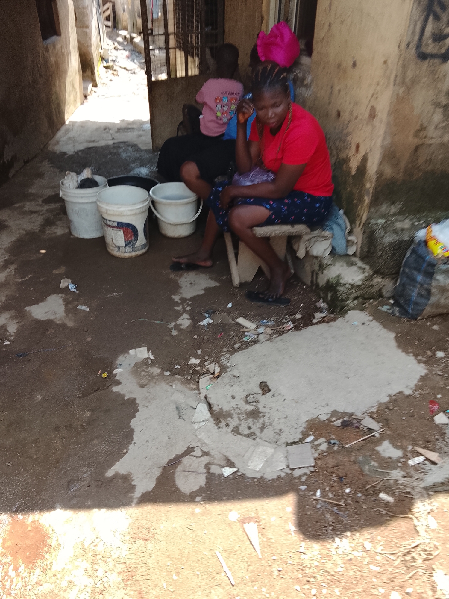 In a narrow, sunlit alleyway in Abuja, Nigeria, a woman with braided hair and a red t-shirt is seated on a wooden stool. She is wearing a dark patterned skirt and black flip-flops. Her left hand rests on her knee, and her right hand is near her face. Beside her, to the left, is another person in a pink t-shirt. In the foreground, three white buckets are placed on the ground. The ground is a mixture of dirt and concrete, with scattered debris. The buildings on either side of the alley are weathered and appear to be made of concrete. Sunlight casts shadows across the scene.