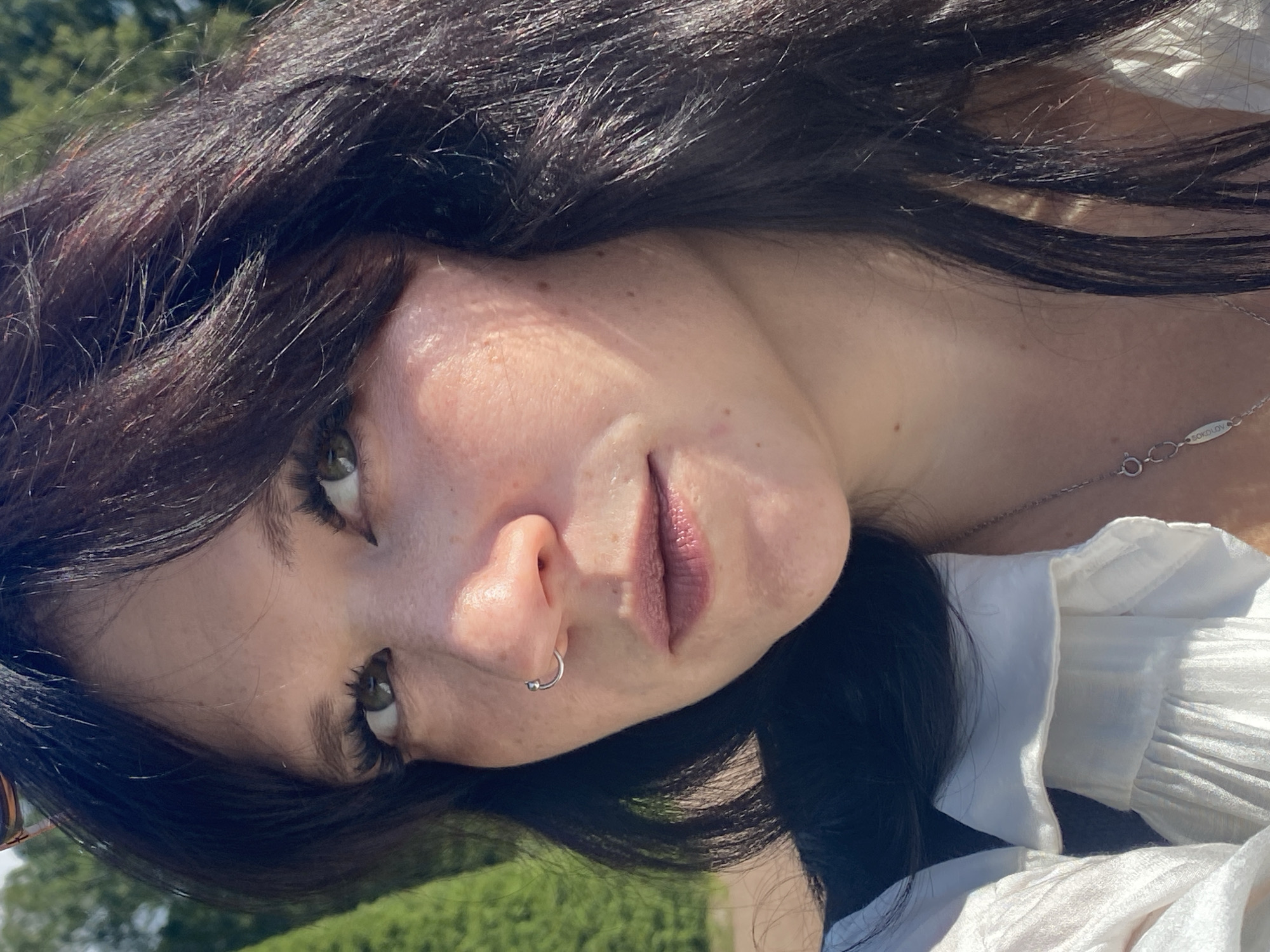 A close-up, outdoor, eye-level medium shot features a woman with dark hair, green eyes, and fair skin. She is wearing a white blouse with ruffles and a silver necklace. A silver nose ring is visible on her left nostril. The background consists of out-of-focus green trees and a bright sky. The woman appears to be looking slightly to her right. The lighting suggests it is daytime, possibly with sunlight casting shadows on her face and hair. The overall scene is relaxed and natural. No specific location within the city of Lomonosov, Russia, can be determined from the visual information provided.