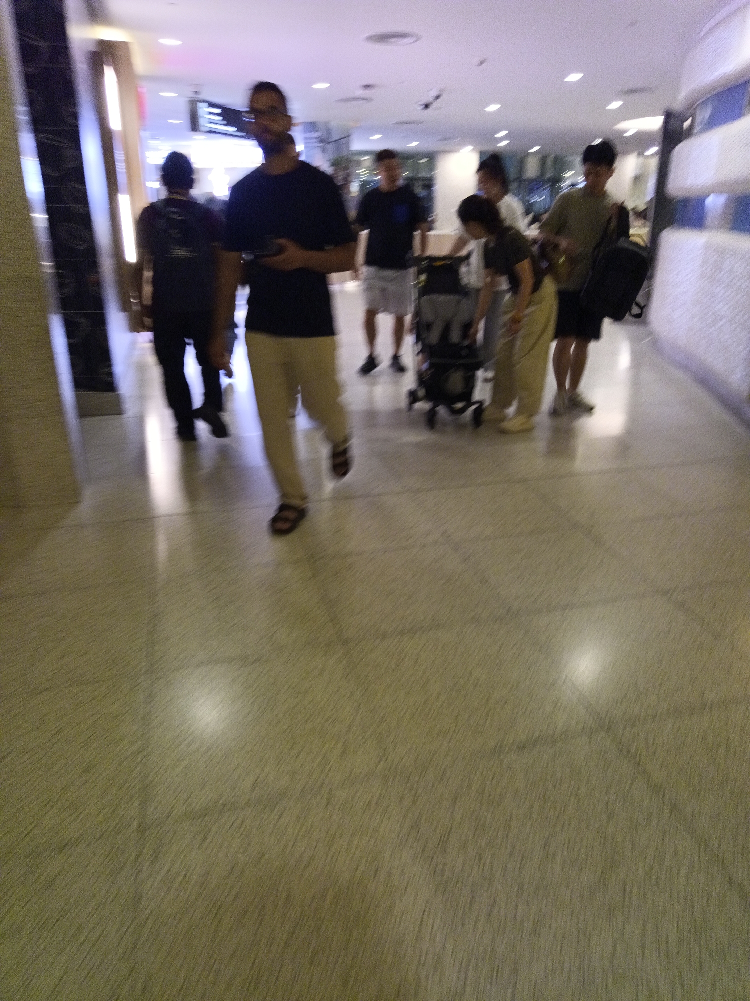 This is a full-body, slightly blurred, candid street photograph taken indoors, likely in an airport or a large shopping mall in Kuala Lumpur, Malaysia. The scene depicts several individuals walking, pushing a stroller, and carrying bags. In the foreground, a man in a dark t-shirt and tan pants is walking away from the camera, holding a phone. To his left, another man in a dark t-shirt and dark pants is also walking away. Further back and to the right, a group of people are walking together. One person is pushing a stroller. The environment appears to be a well-lit corridor with a tiled floor and modern architecture, possibly featuring escalators or elevators on the left. The lighting suggests an indoor environment with artificial lights.