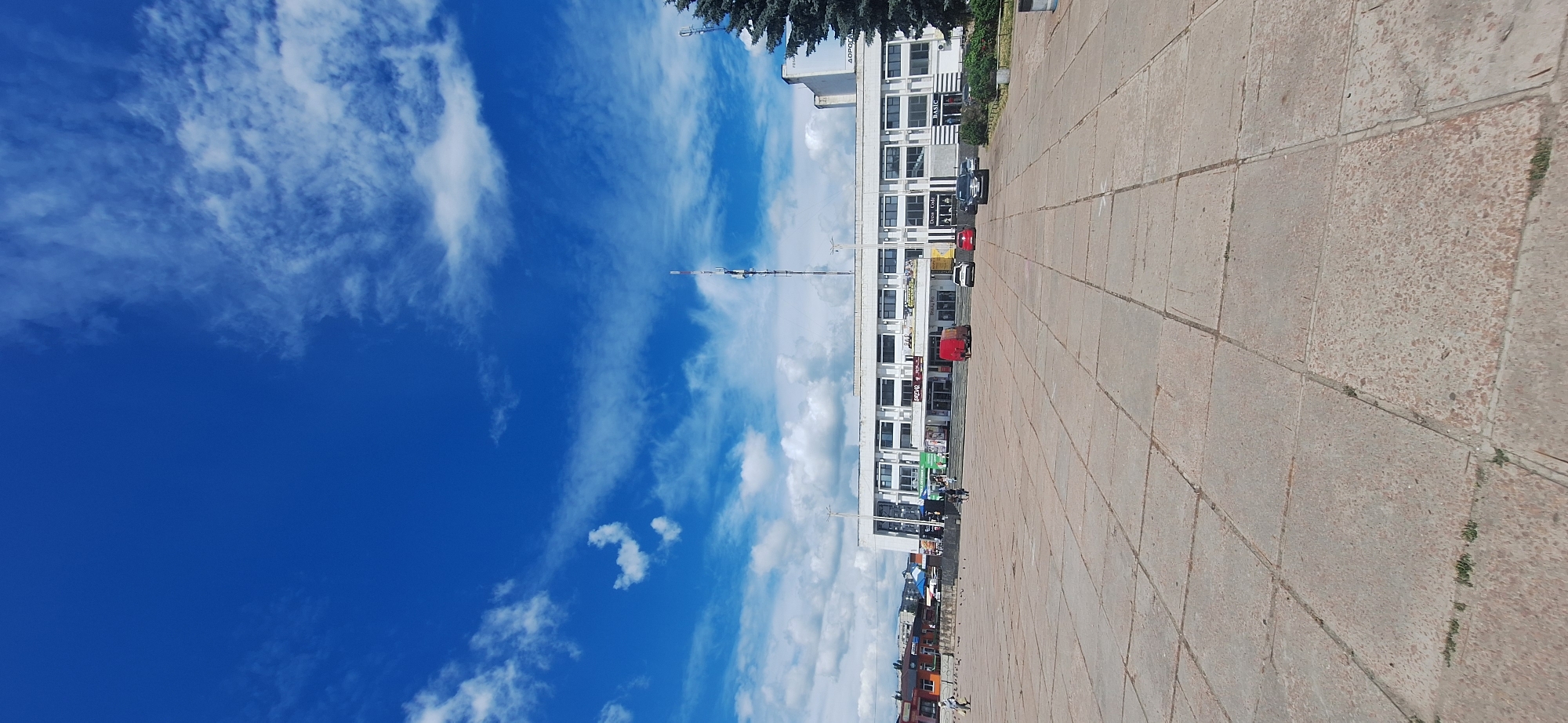 This image captures a wide-angle view of a paved plaza in front of a modern, multi-story building in Bila Tserkva, Ukraine. The sky is a vibrant blue, dotted with fluffy white clouds. The plaza itself is paved with large, square concrete tiles, some of which show signs of wear and small patches of grass growing between them.
In the background, the building is predominantly white with numerous windows and multiple levels. Signs are visible on the building's facade, including one that appears to read "BASIC" and another with Cyrillic text. Several vehicles are parked in front of the building, including a red van, a white car, and a dark SUV. A tall communication tower stands to the right of the building, topped with various antennas and equipment. A large, dark evergreen tree is situated on the far right side of the frame.
Several people are present in the scene, walking on the plaza or near the building entrance. A few pigeons are scattered across the pavement. The overall impression is that of a public square on a clear, sunny day.