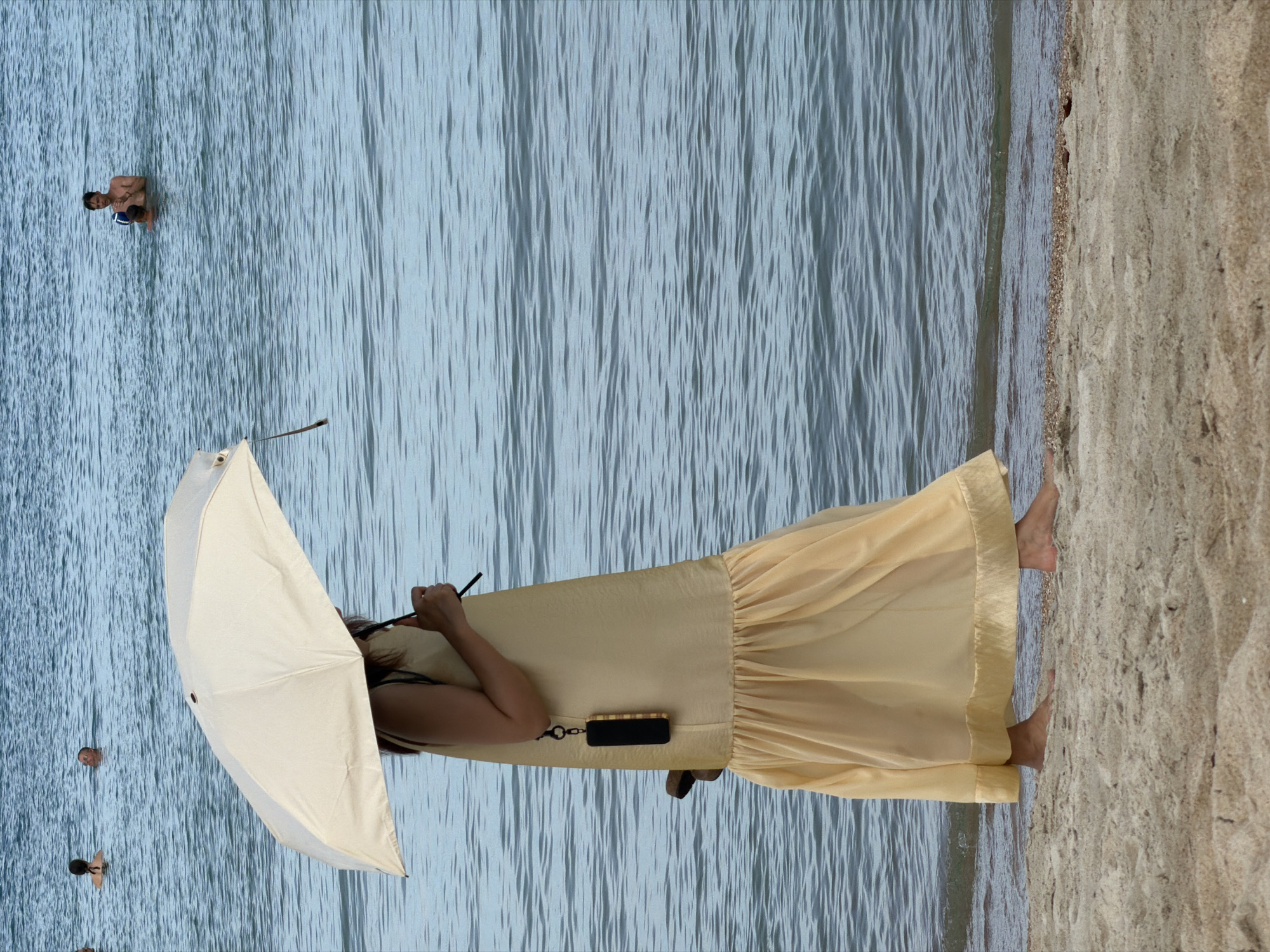 A full-length, side-profile shot shows a woman walking on a sandy beach near the water's edge. She is holding a light-colored umbrella over her head, obscuring her face. She wears a long, flowing yellow dress with a ruffled hem and a dark strap over her shoulder. A black smartphone case hangs from a strap attached to her dress. Her bare feet are visible on the sand. In the shallow water behind her, three other people are present. One person appears to be partially submerged, another is standing in the water, and a third, a male, is also standing in the water holding a dark object. The water is a deep blue with visible ripples. The beach sand is light brown and granular. The sky is not visible in this frame. The location is stated to be Baan Chaweng, Thailand.