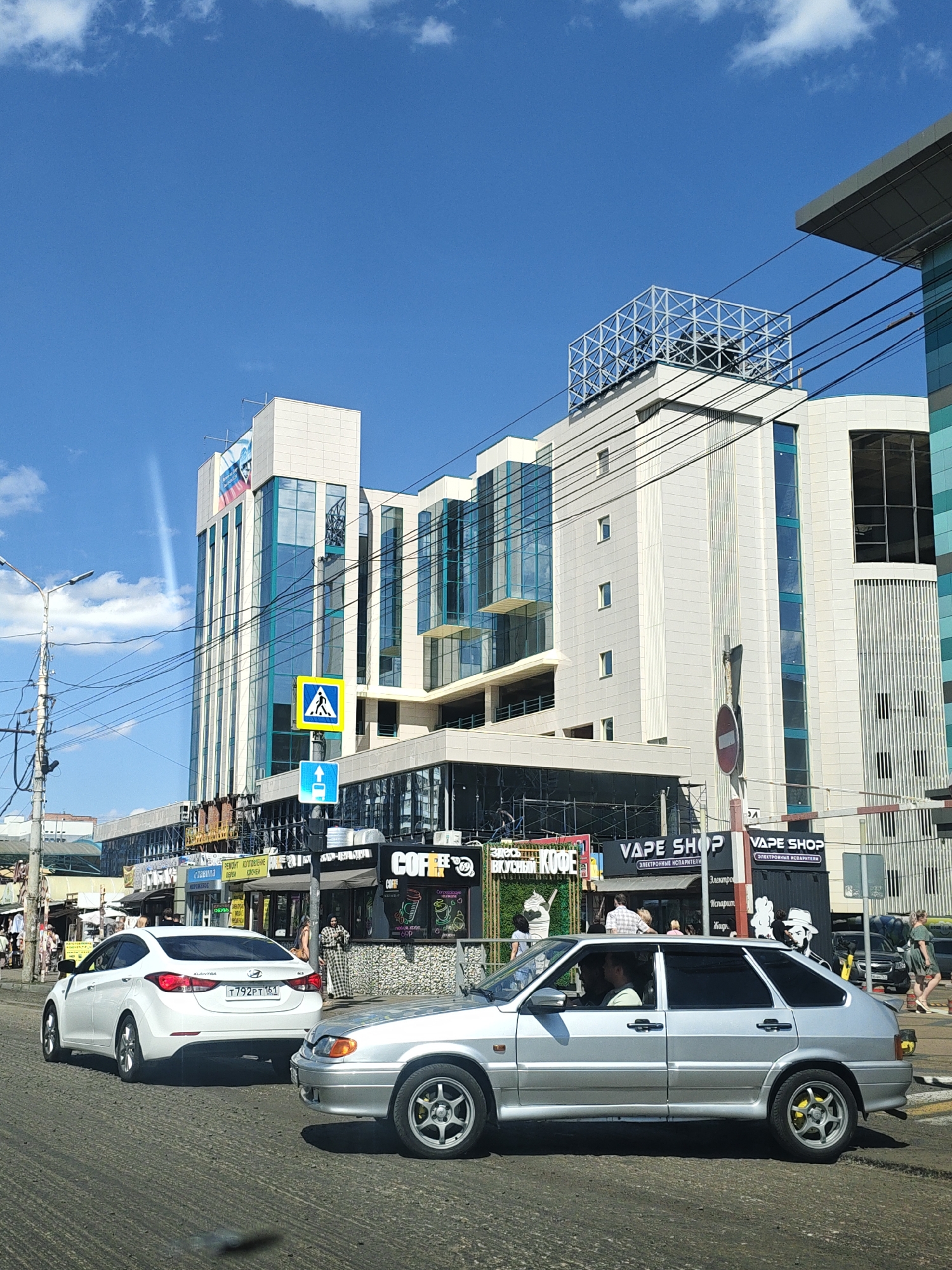A street scene in Rostov-on-Don, Russia, on a sunny day with a clear blue sky and scattered clouds. The foreground features a paved road with two cars, a silver sedan and a white sedan. The silver sedan, a Lada, is parked on the right side of the road, facing left, with its front visible. The white sedan, a Hyundai Elantra, is in the left lane, facing left, with its rear and side clearly visible. Its license plate reads "T 792 PT 161".
In the middle ground, there are several shops and storefronts lining the street. Signs for "VAPE SHOP" and "COFFEE" are prominently displayed. People are seen walking and standing in front of these establishments. One sign above a building displays the Russian flag.
The background is dominated by a large, modern multi-story building with a white facade and glass windows. It appears to be an office building or shopping center. There are power lines and streetlights in the mid-ground. The overall environment suggests an urban setting.