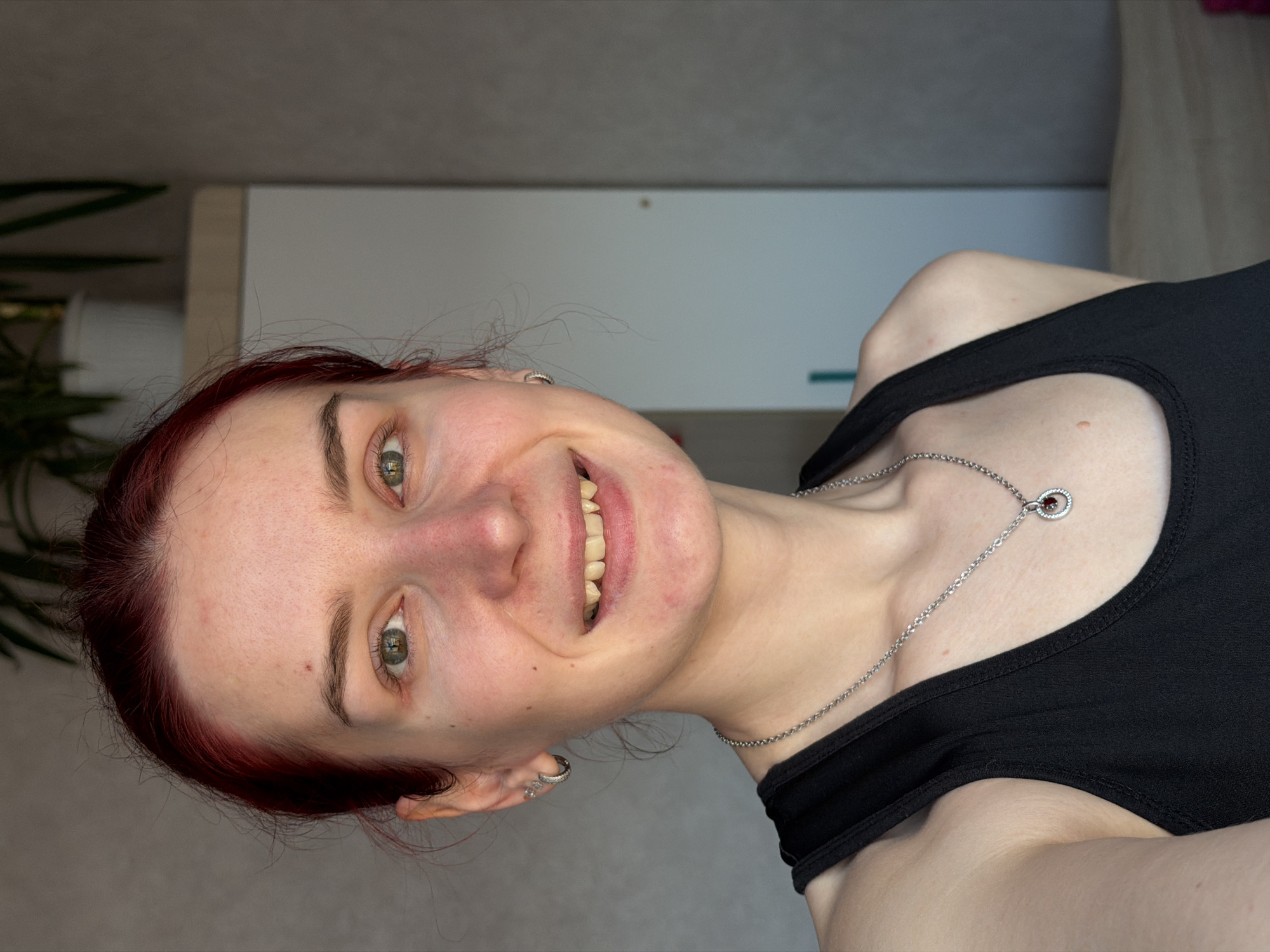 A fair-skinned person with reddish hair, likely female, is smiling and looking at the camera. She is wearing a black tank top and a silver necklace with a circular pendant. The setting appears to be indoors, with a white cabinet or door visible in the background. A green plant is slightly visible in the upper left corner. The location in Chaikovsky, Russia is not discernible from the image.