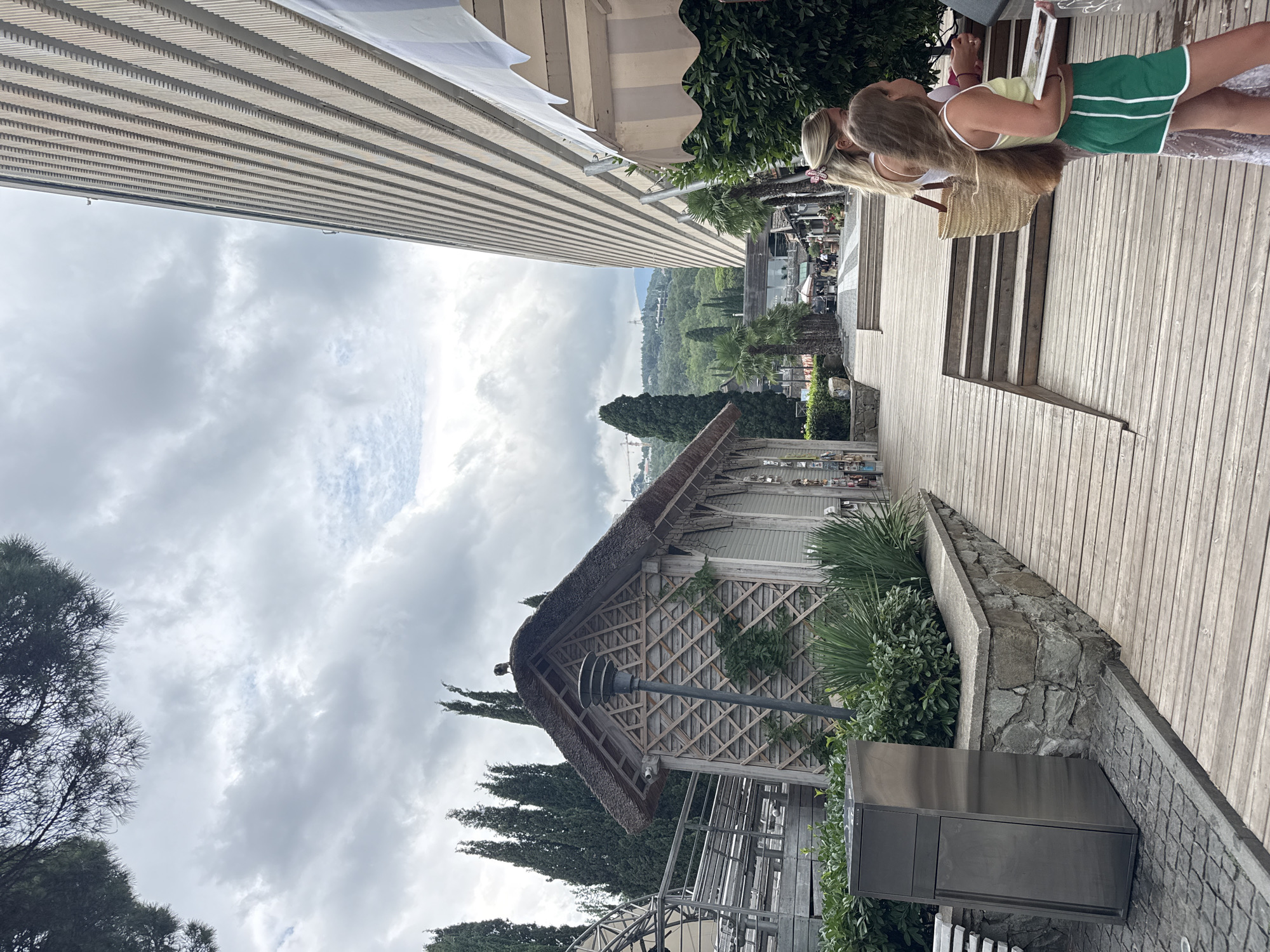 The image captures an outdoor scene in Yalta, Ukraine, featuring a pathway made of wooden planks leading past a thatched-roof wooden building with a lattice facade. In the foreground, a modern stainless steel trash receptacle stands on a cobblestone area. To the right, two females are present; one, an adult with blonde hair, carries a straw bag and stands near a young girl who is holding a thin, rectangular object, possibly a book or tablet. The girl is wearing a yellow tank top and green shorts. Behind them, a large, modern building with a ribbed facade dominates the background, juxtaposed with a cloudy sky. Further back, cypress trees and lush greenery are visible, with hills and mountains in the distance. The overall atmosphere suggests a daytime scene in a resort or promenade area.