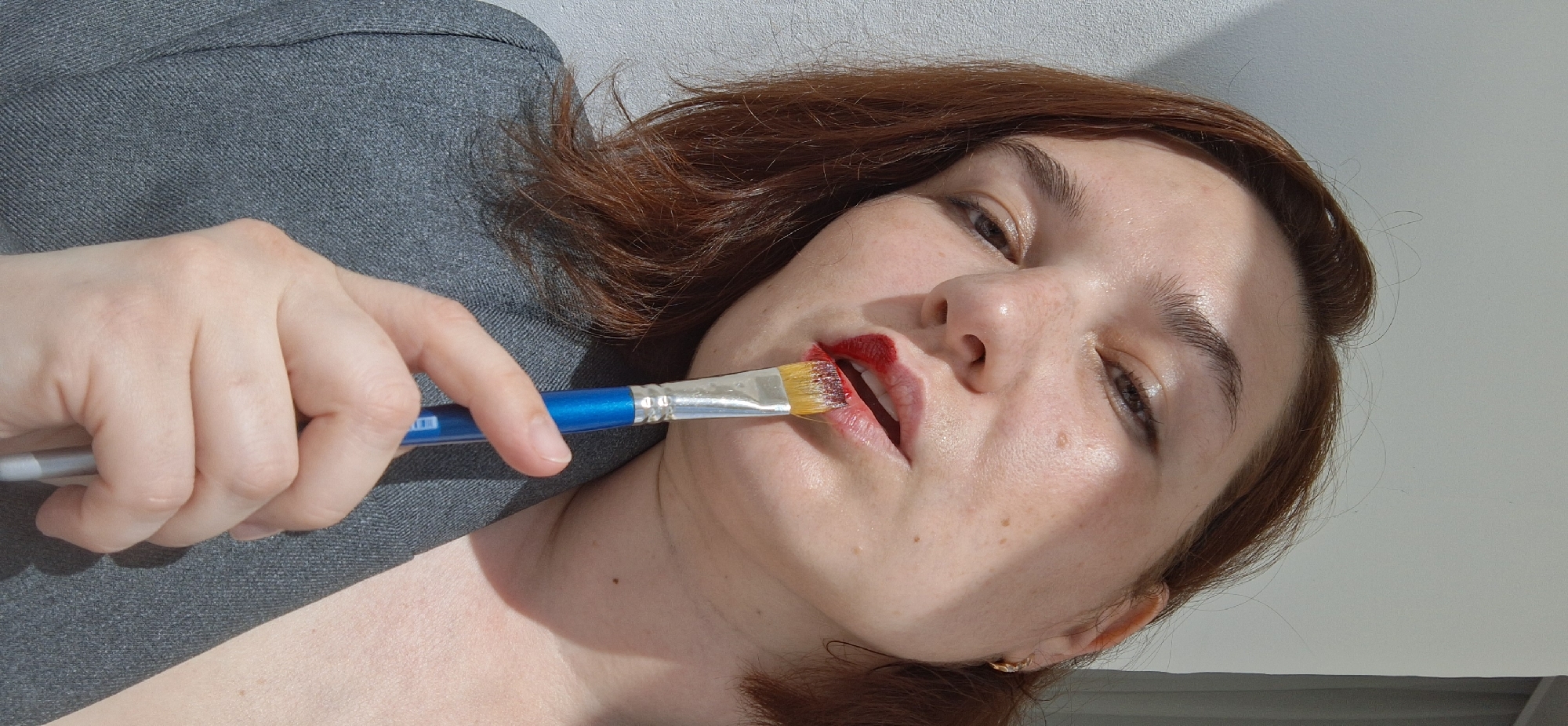 A woman with auburn hair, wearing a gray blazer, is applying red lipstick to her lips using a blue-handled paint brush. The scene appears to be indoors with a light-colored wall in the background. The location is Chaikovsky, Russia.