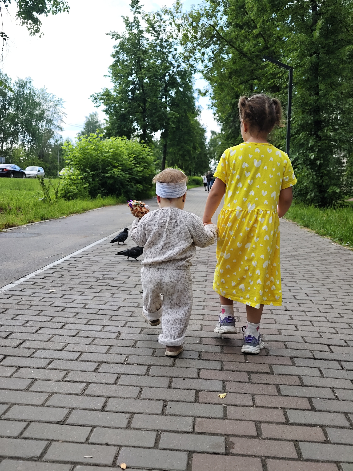 Two young children are walking away from the camera down a brick-paved pathway in Chaikovsky, Russia. The child on the left, wearing a light-colored, patterned jumpsuit and a white headband, holds a toy or object in their right hand, with two pigeons near their feet. The child on the right wears a bright yellow dress with white hearts and purple and white sneakers. They are holding hands. The pathway is bordered by a grassy area and a paved road with parked cars in the background. Lush green trees line the pathway and the background. The sky is overcast.