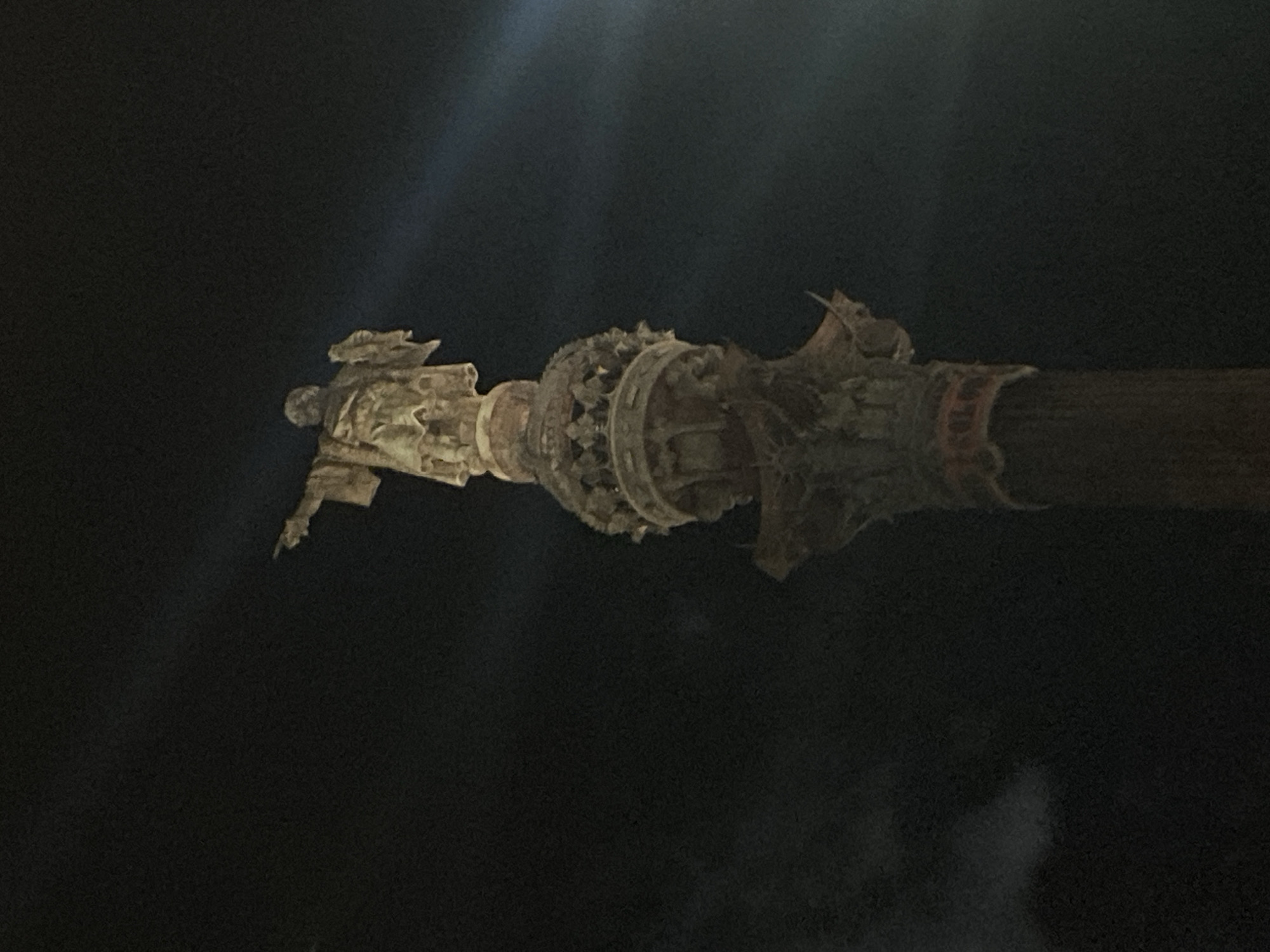 A nighttime exterior shot shows the top portion of the Christopher Columbus Monument in Barcelona, Spain. The monument, a tall column topped by a statue of Columbus, is illuminated from below, creating a dramatic effect against the dark sky. The statue depicts Columbus with his right arm extended, pointing eastward. The column itself is intricately decorated with classical architectural elements.