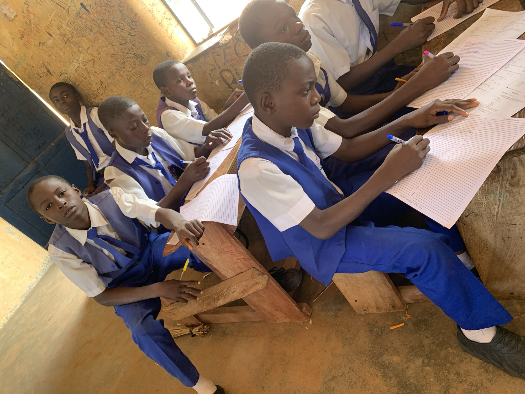 In Akwanga, Nigeria, several young male students, all wearing matching blue and white school uniforms, are seated at rough wooden desks, diligently working on papers. The room appears to be a classroom with dirt floors and a wall covered in graffiti. The students are actively engaged in writing, with pens in hand, focused on their assignments. Some students are looking down at their papers, while others glance up, perhaps contemplating their work. The atmosphere suggests a typical school day, with an emphasis on education and learning.