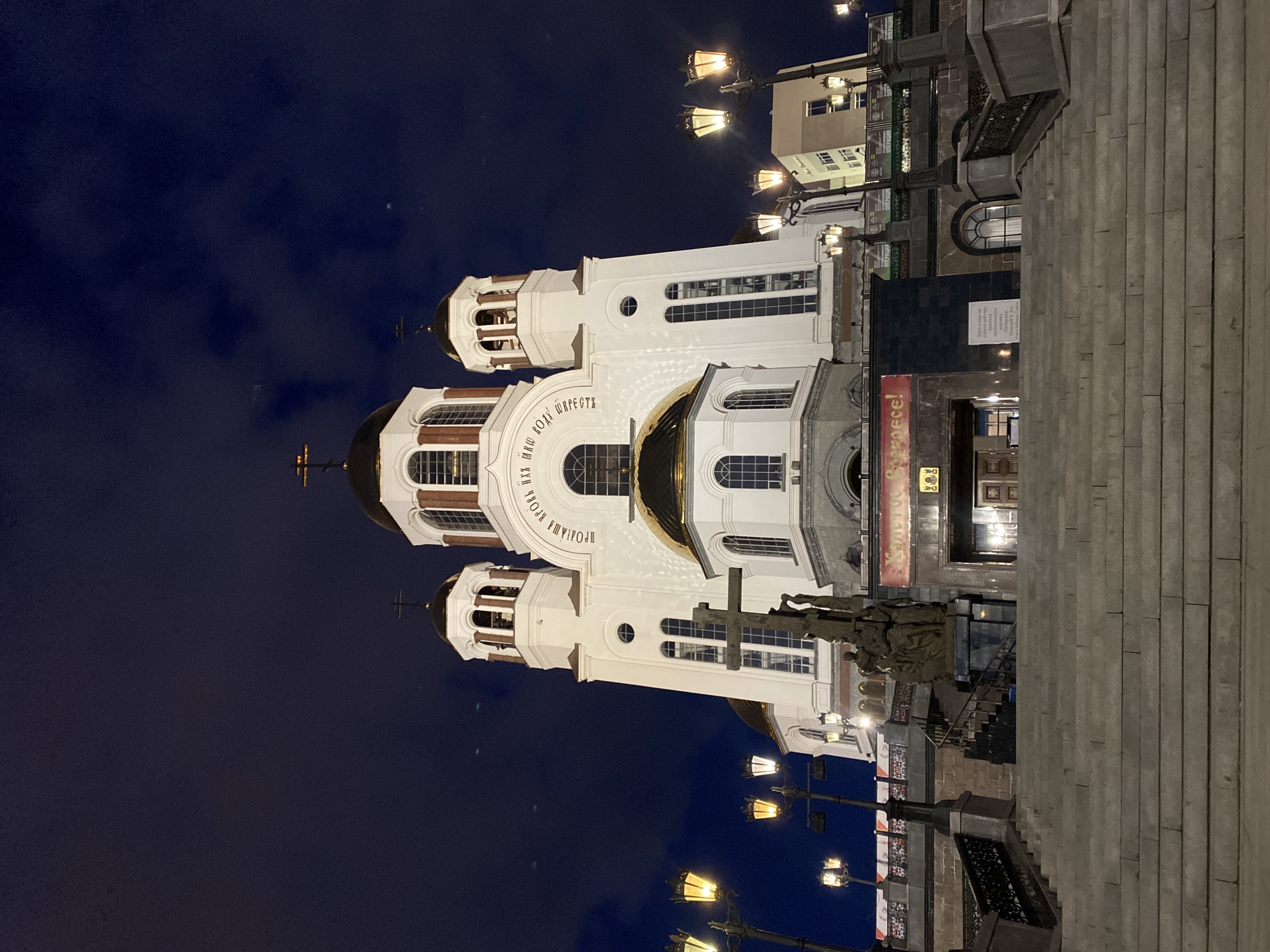 At night, the Church on the Blood in Yekaterinburg, Russia, is seen from ground level. The white church with gold domes is brightly illuminated against a dark blue sky. Street lamps are visible in the foreground, and steps lead up to the church entrance. A bronze statue is present near the church. A multi-story building stands next to the church on the right.