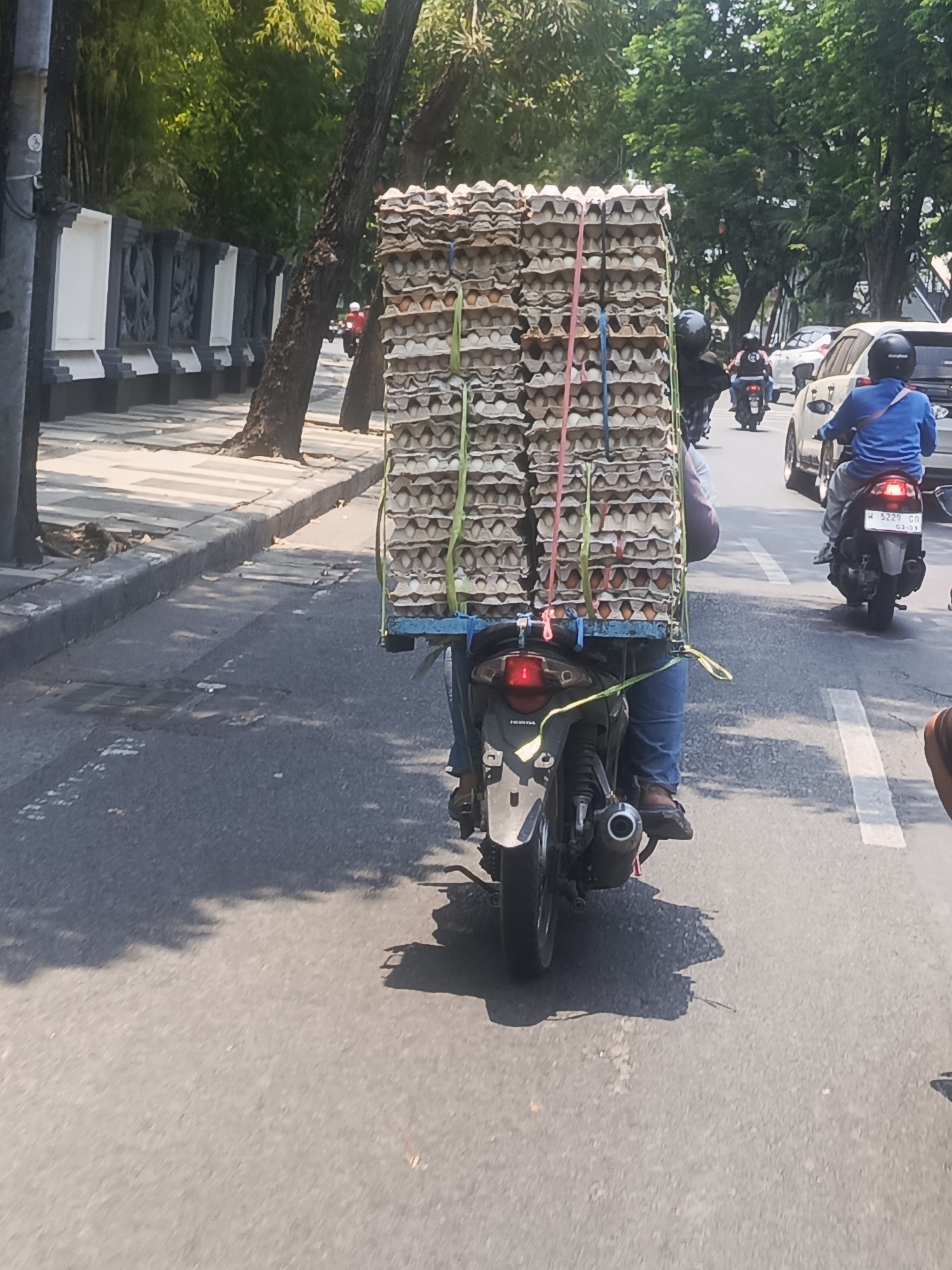 A motorcycle is carrying a large stack of egg cartons on a street in Surabaya, Indonesia. The motorcycle is dark-colored and the egg cartons are stacked high, secured with ropes. The rider is wearing a blue shirt and blue jeans. In the background, other vehicles, including cars and motorcycles, are visible, as is a sidewalk with trees and a decorative wall. The sun is shining, casting shadows on the road.