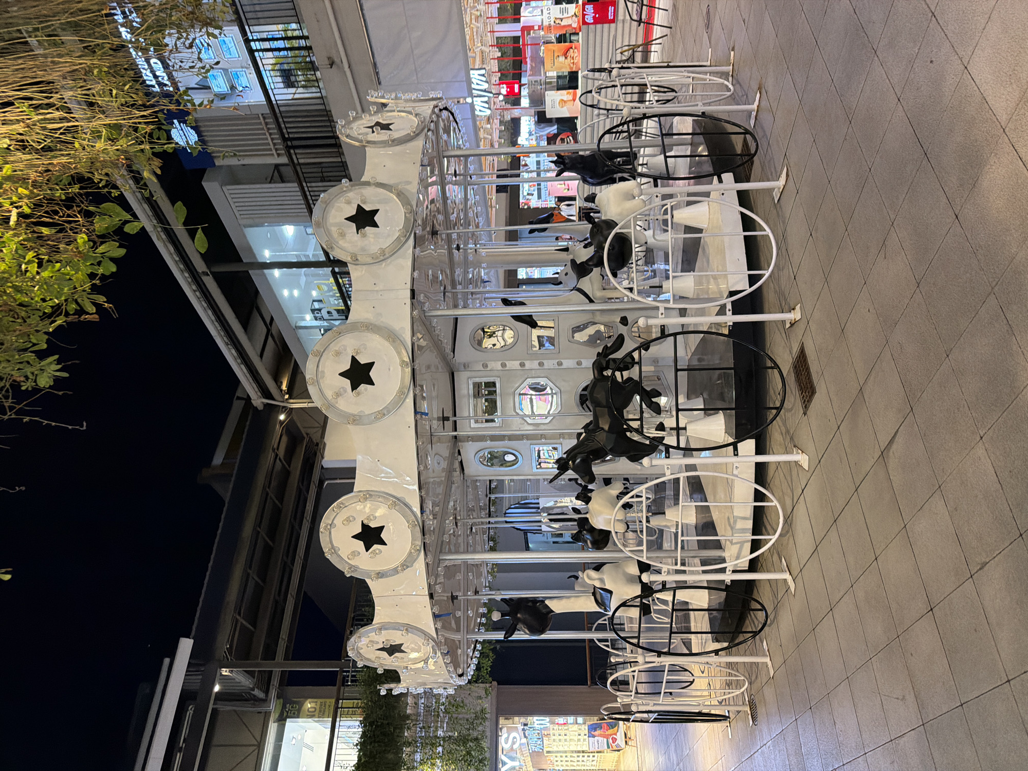 This media file depicts an outdoor scene at night in Baan Chaweng, Thailand. The primary subject is a carousel-like structure with black and white cow figures. The carousel is decorated with circular elements featuring stars and lights. It is a nighttime shot, indicated by the dark sky and artificial lighting illuminating the scene.
The carousel appears to be in a pedestrianized area or walkway, with tiled pavement visible in the foreground. In the background, there are buildings with illuminated storefronts, suggesting a commercial or entertainment district. Some of the storefronts have visible text, though it is mostly illegible due to the angle and lighting. One sign appears to read "YS," and another partially visible sign has Cyrillic text. A tree with green foliage is visible on the left side of the frame, adding a natural element to the urban setting. The overall mood is festive and illuminated, typical of a lively evening in a tourist area. No people are clearly visible in the frame.
