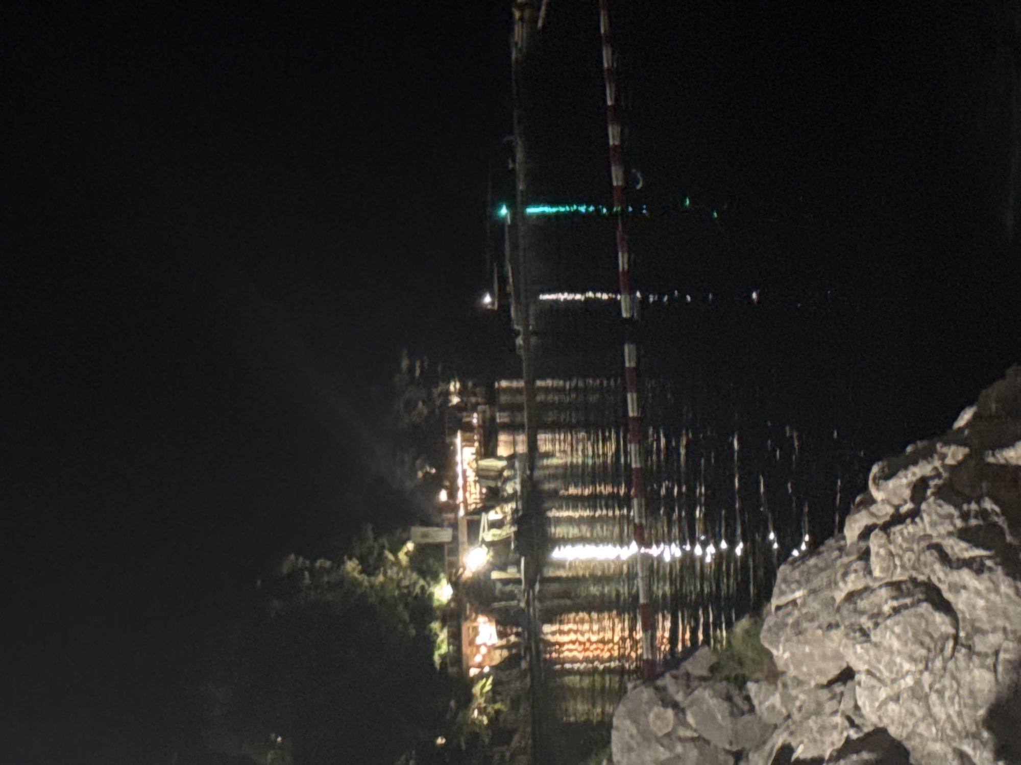 The image depicts a nighttime scene of a waterfront location, possibly a beach or a harbor area, in the city of Budva, Montenegro. In the foreground, large rocks are visible on the left side. The water in the middle ground reflects the lights from buildings on the shore, which appear to be illuminated with warm, yellow and orange tones. These buildings are situated along a tree-lined embankment. In the water, a red and white striped barrier is visible, possibly marking a swimming area or a boundary. Further in the background, on the right side of the water, a green light is emanating from a structure or a boat. The overall atmosphere is dark and illuminated by artificial lights.