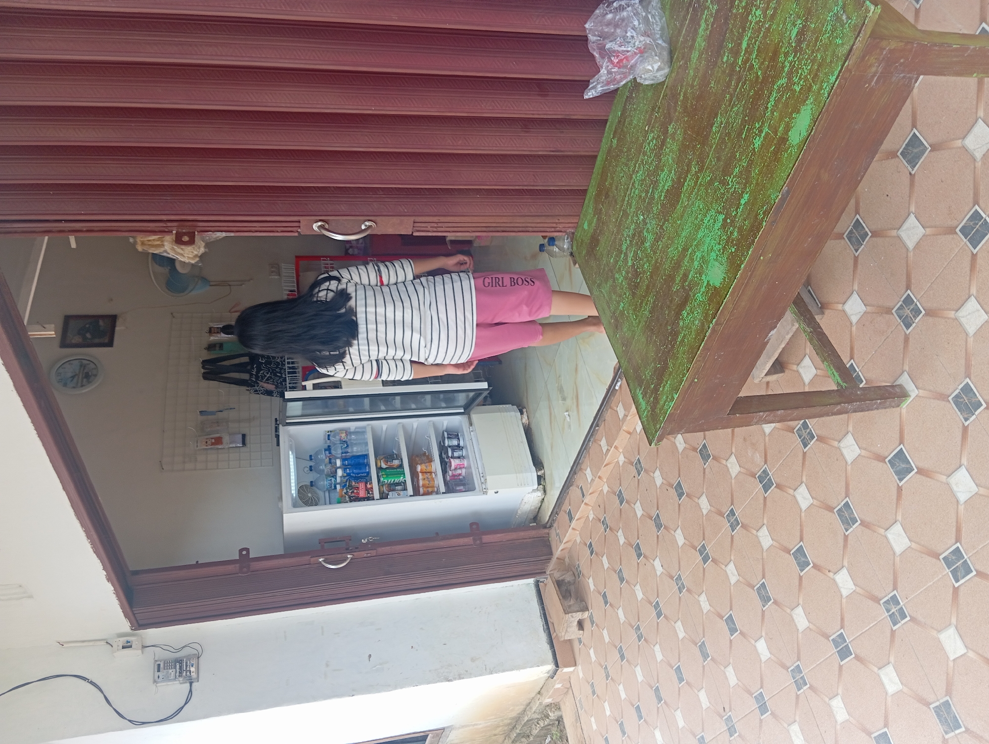 A young Indonesian female, facing away from the camera, stands at the entrance of a small shop in Rejosari, Indonesia. She is wearing a striped long-sleeved shirt and pink shorts with the text "GIRL BOSS" visible on the left leg. The shop, open to the outdoors, features a white refrigerator stocked with drinks, and a metal grate shelf displaying various items. Adjacent to the entrance, a red corrugated metal folding door is partially open. Outside the shop, on a tiled patio, a weathered green wooden table with a plastic bag containing a red object sits in the foreground. A power meter is mounted on the white exterior wall to the left of the entrance.