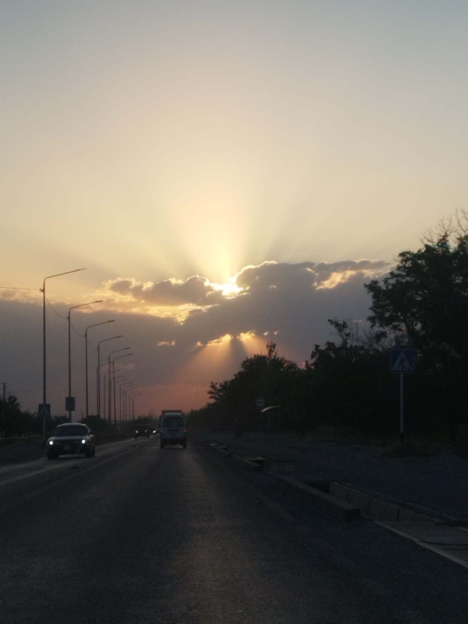 The image captures a dusky highway scene in Shymkent, Kazakhstan, with the sun setting dramatically behind clouds. The sky is a gradient of pale yellow to light blue, with distinct crepuscular rays emanating from behind a band of dark clouds. A line of streetlights runs along the left side of the road, diminishing in the distance. Several vehicles are visible on the asphalt road: a white van in the middle lane, a dark sedan approaching in the left lane with its headlights on, and other vehicles further ahead. Trees and foliage frame the right side of the road, obscuring much of the background. A pedestrian crossing sign with a blue square and white pedestrian symbol is visible on the right side of the road, near a streetlight. The overall impression is one of a tranquil end to the day on a busy road.