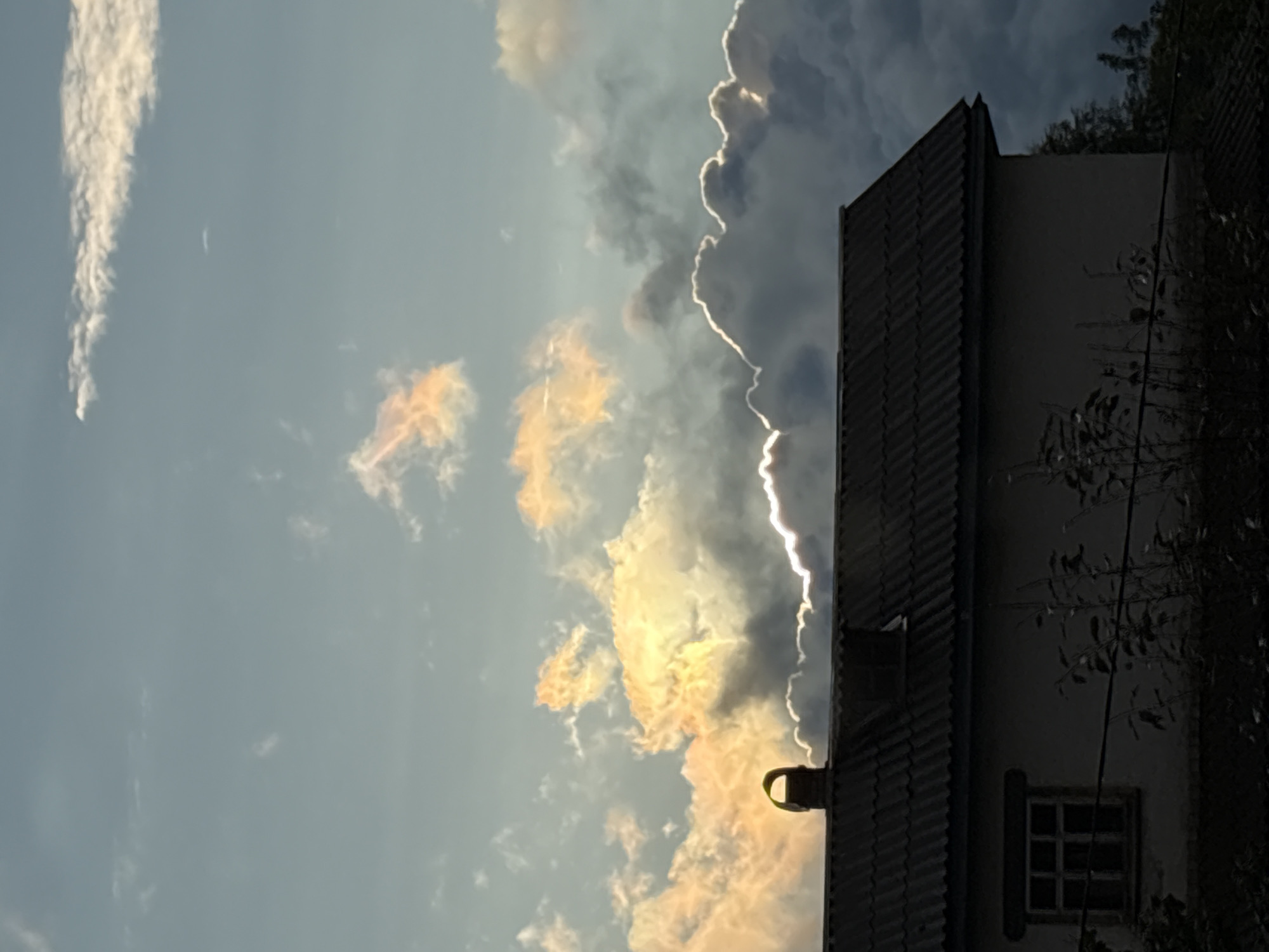 The image captures a twilight sky over a house in Vetluzhsky, Russia. The sky is filled with dramatic clouds, some tinged with the warm glow of the setting sun, while others are dark and ominous. The upper part of the sky is a pale blue, fading into lighter hues as it meets the horizon.
In the foreground, the dark silhouette of a house is visible. It features a gray, corrugated roof, with a chimney standing prominently on the left side. A single, dark-framed window is visible on the left wall of the house. The lower part of the image is shrouded in shadow, with the dark shapes of vegetation and possibly a fence line. The overall impression is one of a quiet, slightly melancholic evening in a rural or suburban setting.
