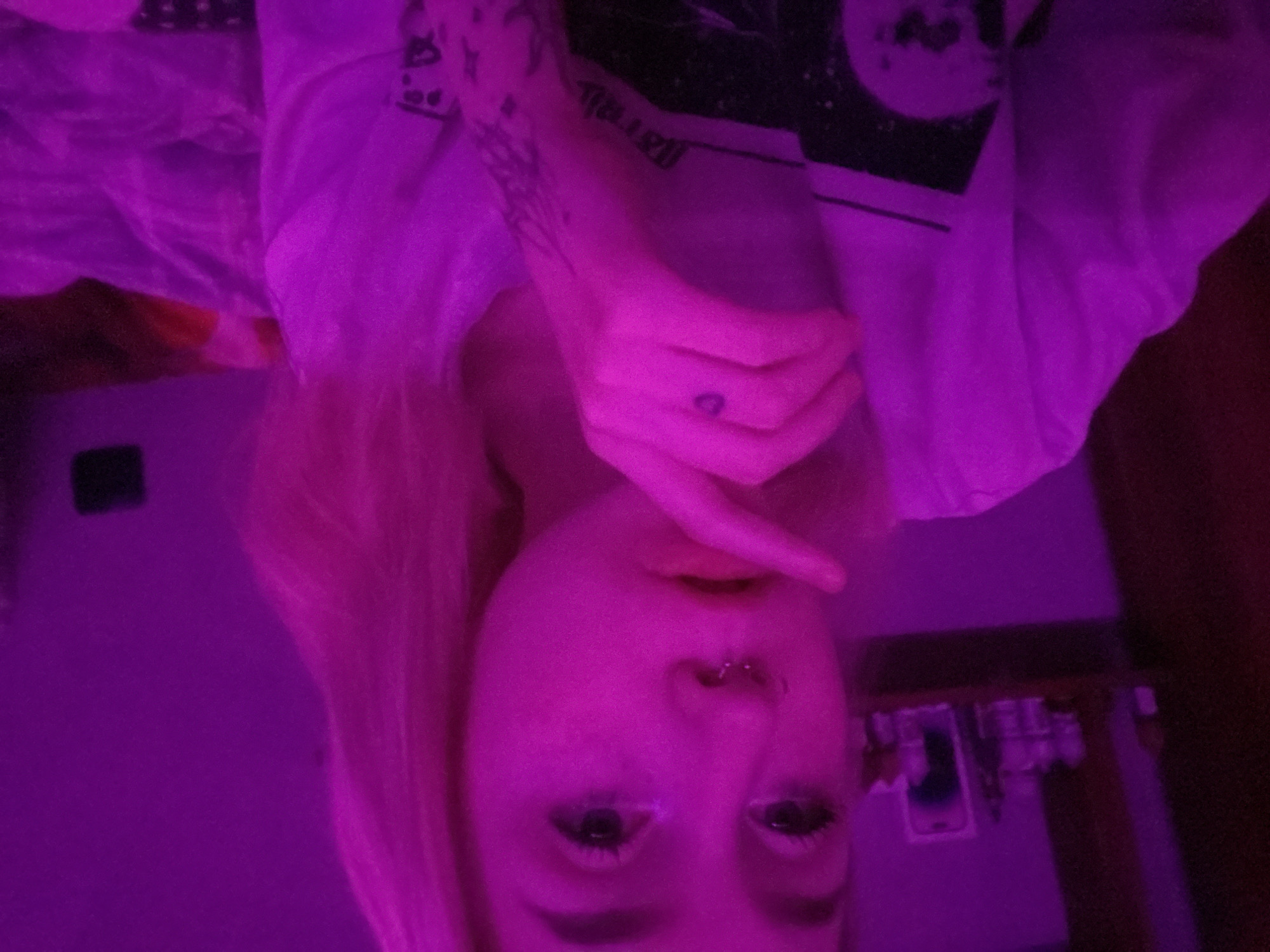 The image presents a close-up indoor portrait of a young individual, likely female, captured under dominant purple-magenta lighting. The subject has light-colored hair and a visible septum piercing. The right hand is raised, with the index finger placed against the lips. A small, dark circular tattoo is visible on the tip of the right index finger, and multiple dark-inked tattoos are present on the left forearm. The subject is wearing a light-colored (appearing white under the lighting) short-sleeved t-shirt featuring a dark graphic design with geometric and textual elements on the chest area. The background, blurred due to shallow depth of field, shows a wall. To the subject's left, a dark shelf or surface holds several indistinct objects, appearing as bottles, jars, and a standing rectangular item, possibly a phone or framed picture. To the subject's right, a portion of a bed or similar furniture covered with a patterned blanket is visible. The specific location within the city of Izhevsk, Russia, cannot be determined from the visual evidence presented in the image.