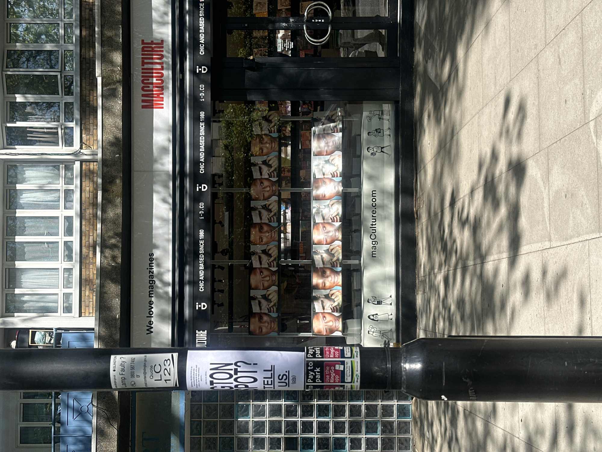 The image shows the storefront of MagCulture, a magazine shop in London, United Kingdom. The building has a white façade with the company name in red. The shop window displays magazine covers, with the text "CHIC AND BASED SINCE 1980 i-D.CO" and "magCulture.com". There is a glass door with metal handles, indicating the entrance. A black metal lamp post stands in front of the store with stickers and signs on it, including details about reporting faulty lamps and paying for parking. The street outside appears paved, with shadows cast across it. The architecture in the background includes brick and windows.