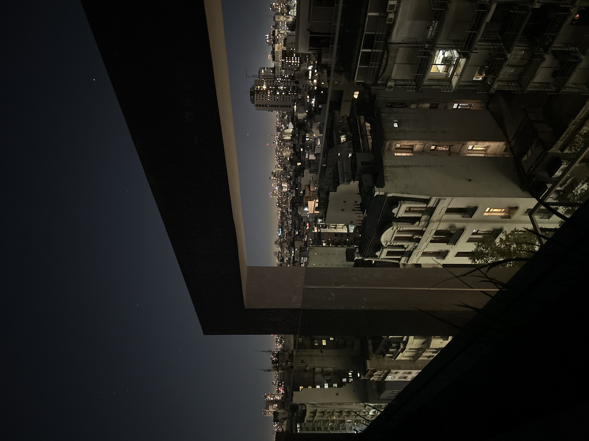 From an elevated position, the image captures an urban landscape at night, possibly from a balcony or rooftop in Buenos Aires, Argentina. The cityscape stretches horizontally, with distant high-rise buildings illuminated by numerous lights against a dark sky. Closer to the viewer are structures of varying heights, some in shadows, with lit windows indicating occupancy. Architectural details are partially visible, including balconies and building facades. A section of a concrete structure is present in the foreground. The sky shows faint stars.