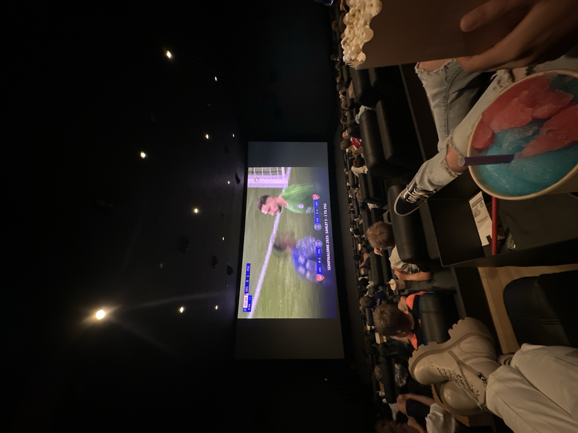 The image captures a scene inside a movie theater, presumably in Horsens, Denmark. The focus is on the audience watching a soccer game being projected onto the large screen. The game being displayed is "SEMIFINALERNE 2025: SAMLET 3-1 TIL PSG" with the score being ARS 0-1 PSG during the 45th minute. Viewers, including some children, are seated in rows of theater seats. An individual is holding a beverage with a blue and red ice slush and a container of popcorn. The theater lighting is dim, with some ceiling lights visible.