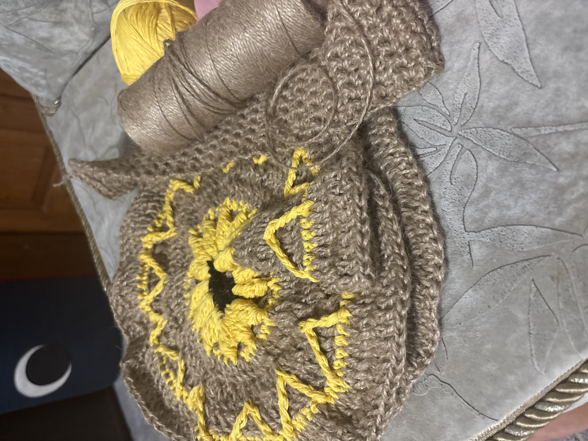 The image shows a close-up view of a crochet project, likely a sunflower motif, made with brown and yellow yarn. The project is laid out on a textured surface that appears to be a tablecloth or some other fabric with a floral pattern. To the right of the crochet work is a spool of brown twine and a ball of bright yellow yarn. In the background, a dark blue object with a white crescent moon symbol is visible. The overall scene suggests a crafting or hobby activity. There is no indication of the city of Zmiiv, Ukraine, or any specific urban environment.