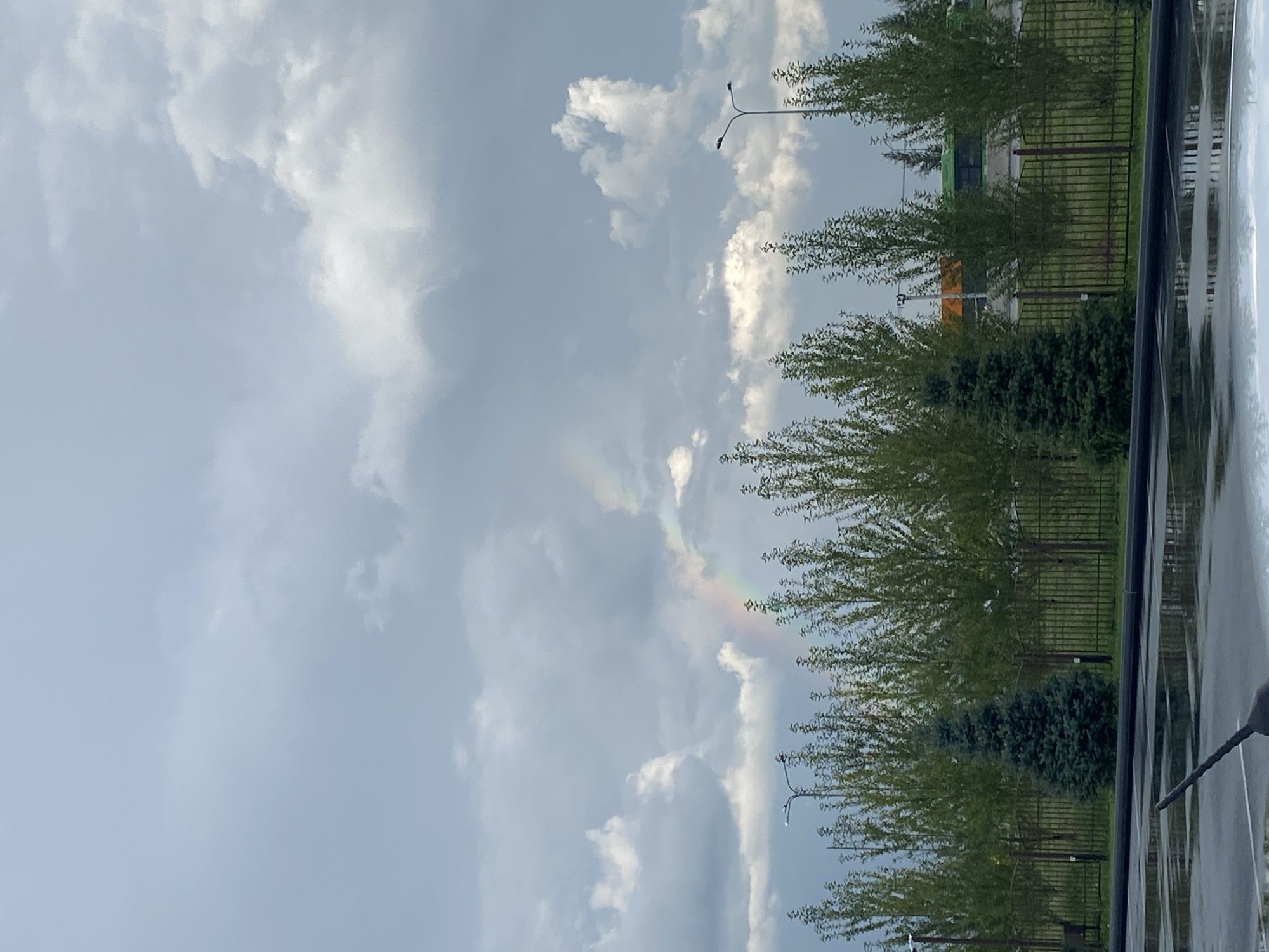 The image shows a cloudy sky with a faint rainbow visible. In the foreground, there is a wet asphalt surface reflecting the sky and trees. A dark object, possibly a broom or a part of a vehicle, is partially visible in the bottom left corner. Beyond the asphalt, there is a black metal fence with green trees and some smaller evergreen trees behind it. In the background, there are streetlights and what appear to be buses or other vehicles, suggesting an urban or suburban setting. The scene suggests a recent rain shower, given the wet surfaces and the presence of a rainbow. The overall impression is that of an outdoor scene in a city, possibly on the outskirts, with elements of nature interspersed with urban infrastructure.