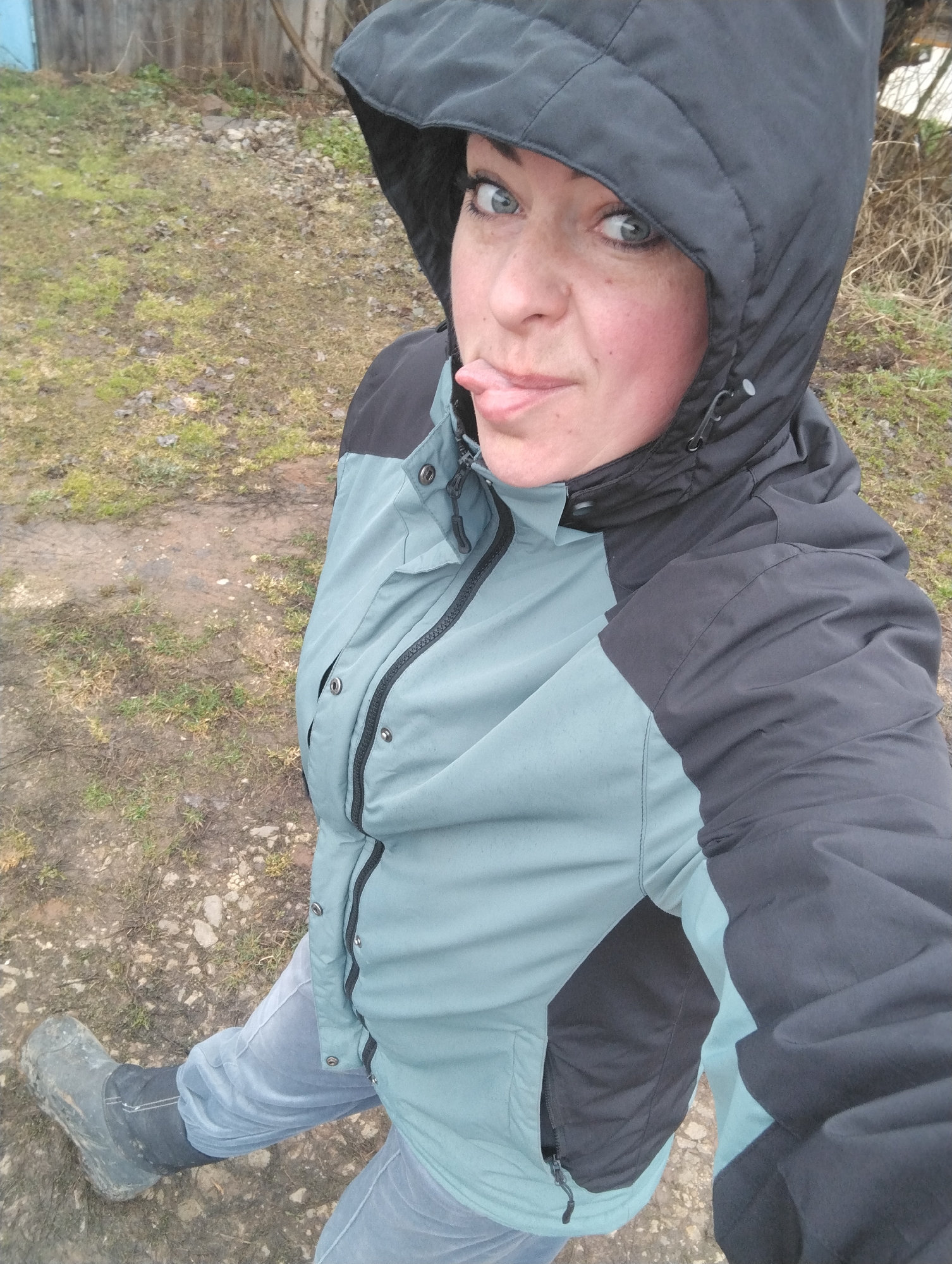 An adult woman is captured in a self-portrait outdoors, seemingly in Grishanki, Russia. She is shown from the waist up, angled slightly from the left, looking directly into the camera. Her expression is playful, with her tongue sticking out to the side, and her blue eyes are wide. She has light skin and some visible freckles on her cheeks.
She is wearing practical, warm clothing suitable for cool or damp weather. Her jacket is two-toned, primarily light blue-green with dark gray or black sections on the shoulders, arms, and hood. The jacket features zippers and snaps. Her hood is pulled up, framing her face. Beneath the jacket, she wears light-colored jeans, and her left foot, which is visible, is clad in a dark, possibly rubber or waterproof, boot that appears muddy.
The background consists of a rustic outdoor setting. The ground beneath her is unpaved, covered with patches of sparse green and brown grass, mud, and small stones. In the distance, behind her, there is a weathered wooden fence and some bare tree branches, reinforcing the rural or rustic location. The lighting suggests it is daytime, possibly overcast, providing soft, diffused light. There is no visible text in the image.