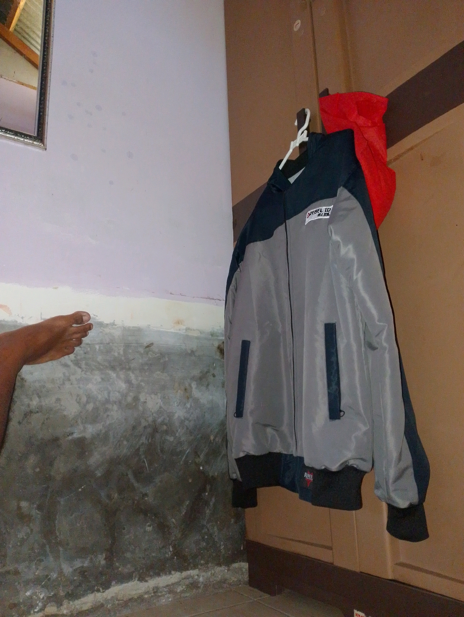 The image depicts an indoor scene, likely a room in a residence in Desa Pagandaran, Indonesia. In the foreground, a gray and dark blue jacket is hanging on a white hanger. The jacket has a red inner lining visible. A label with text, possibly a brand name, is present on the chest of the jacket. To the left of the jacket, a foot is visible, suggesting the presence of a person. The wall behind the jacket appears to be a light purple color, with some darker, rougher textured area below it. To the right of the jacket, a brown wooden surface, possibly a closet door, is visible. The lighting in the scene is dim.