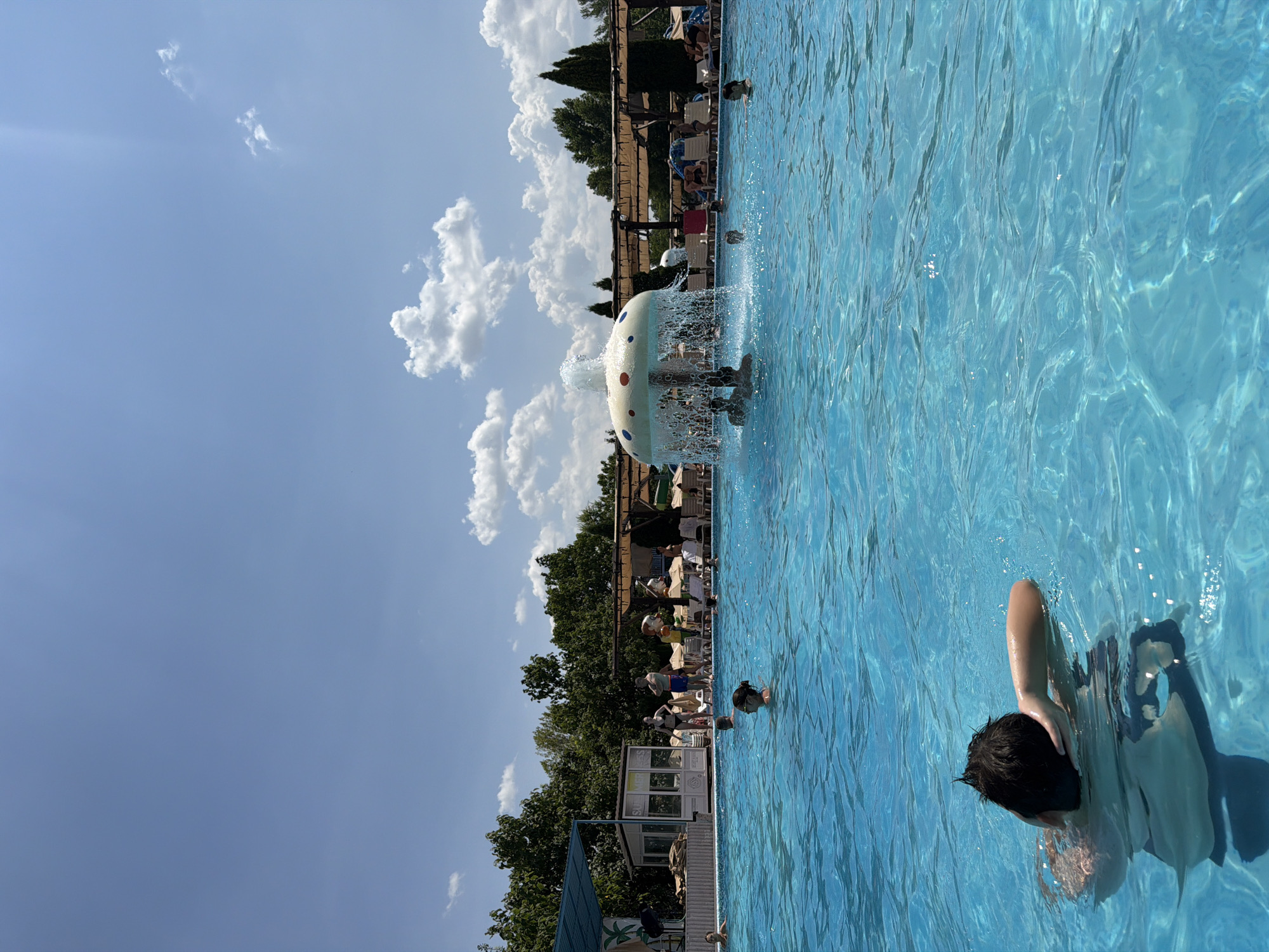 The image depicts a bright, sunny day at a public swimming pool, possibly a water park, in or near the city of Sociteni, Moldova. The pool is filled with clear blue water, with ripples and light reflections on the surface. In the foreground, a young person with dark hair is partially submerged in the water, holding their arm up to their head. Scattered throughout the pool are other individuals, some swimming, others standing or floating.
In the midground, a decorative fountain shaped like a mushroom or toadstool sprays water upwards and outwards, creating a cascade effect. Several people are gathered around the fountain, enjoying the spray.
The background reveals the pool's perimeter with lounge chairs and umbrellas, suggesting an area for relaxation. Beyond the pool, there is a dense line of green trees and vegetation. A building with glass windows and signage that appears to read "SUMMER SES" is visible on the left side of the image. The sky is a clear, bright blue with scattered white cumulus clouds, indicative of fair weather. The overall scene is one of recreation and leisure in an outdoor aquatic environment.