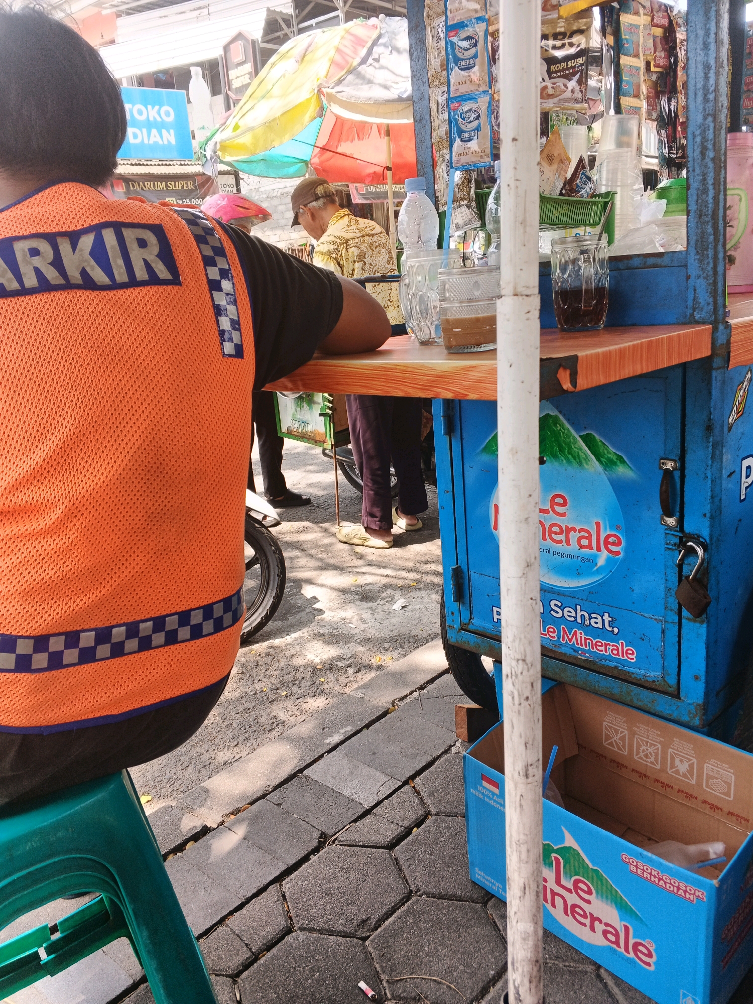 In the city of Kalirejo, Indonesia, a person wearing an orange safety vest with the word "PARKIR" emblazoned on the back is seated on a green plastic stool. The vest has reflective checkered stripes. Behind them, a blue food cart is visible, adorned with promotional materials for "Le Minerale" bottled water. The cart displays imagery of mountains and text indicating "Le Minerale." Various packaged goods and drinks are displayed on shelves on the cart, including coffee sachets and disposable cups. A glass of dark liquid, likely coffee, sits on the cart's wooden countertop next to a bottle of water. In the background, another person in a patterned shirt is seen near a colorful umbrella. The ground is paved with interlocking concrete blocks and some asphalt. A partially opened cardboard box with "Le Minerale" branding is on the ground near the cart. A motorcycle is partially visible behind the person in the safety vest. The scene suggests a street-side vendor operation.