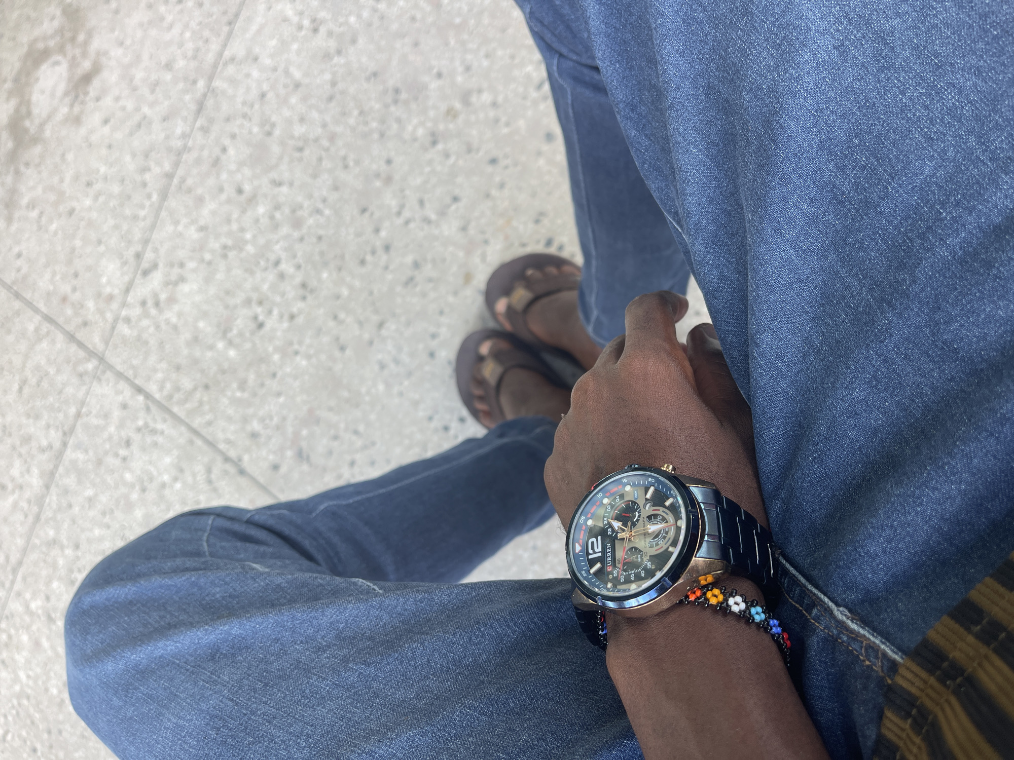 An overhead, high-angle shot captures a person sitting, looking down at their lap and feet on a light-colored, speckled ground, possibly in Dar es-Salaam, Tanzania. The subject's dark-skinned hands rest on their blue jeans, which are slightly faded.
On the left wrist, a prominent black and rose-gold chronograph watch with multiple dials is visible. The brand "CURREN" is clearly displayed on the watch face, along with a large numeral "12" and smaller markers like "20," "40," and "60," indicating a stopwatch function ("CHRONOGRAPH"). Below the watch, a colorful beaded bracelet, featuring small white, yellow, orange, red, green, and blue beads, encircles the wrist.
The person is wearing dark brown, open-toed sandals, with only the upper parts of the feet visible. A glimpse of a striped garment, possibly a shirt or jacket in brown and yellow tones, can be seen beneath the jeans on the right side of the frame. The floor appears to be terrazzo or a similar material, with faint lines suggesting tiling or cracks. The lighting suggests it is daytime.