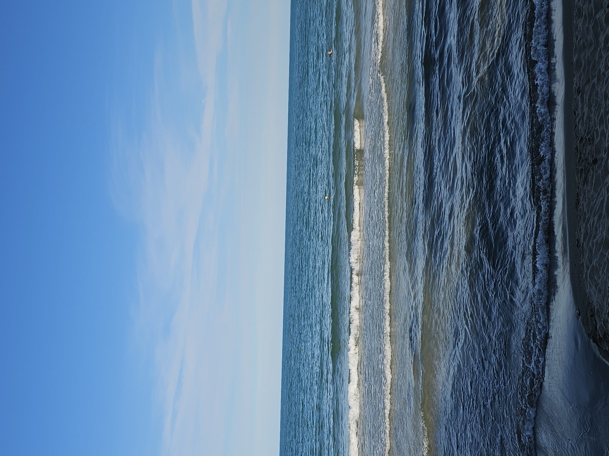 The image shows a beach scene with the ocean and sky. The sky is a clear blue with some wispy white clouds. The ocean is a deep blue with gentle waves rolling towards the shore. There is a sandy beach in the foreground. Two yellow buoys are visible in the water, and a person is swimming in the distance.
