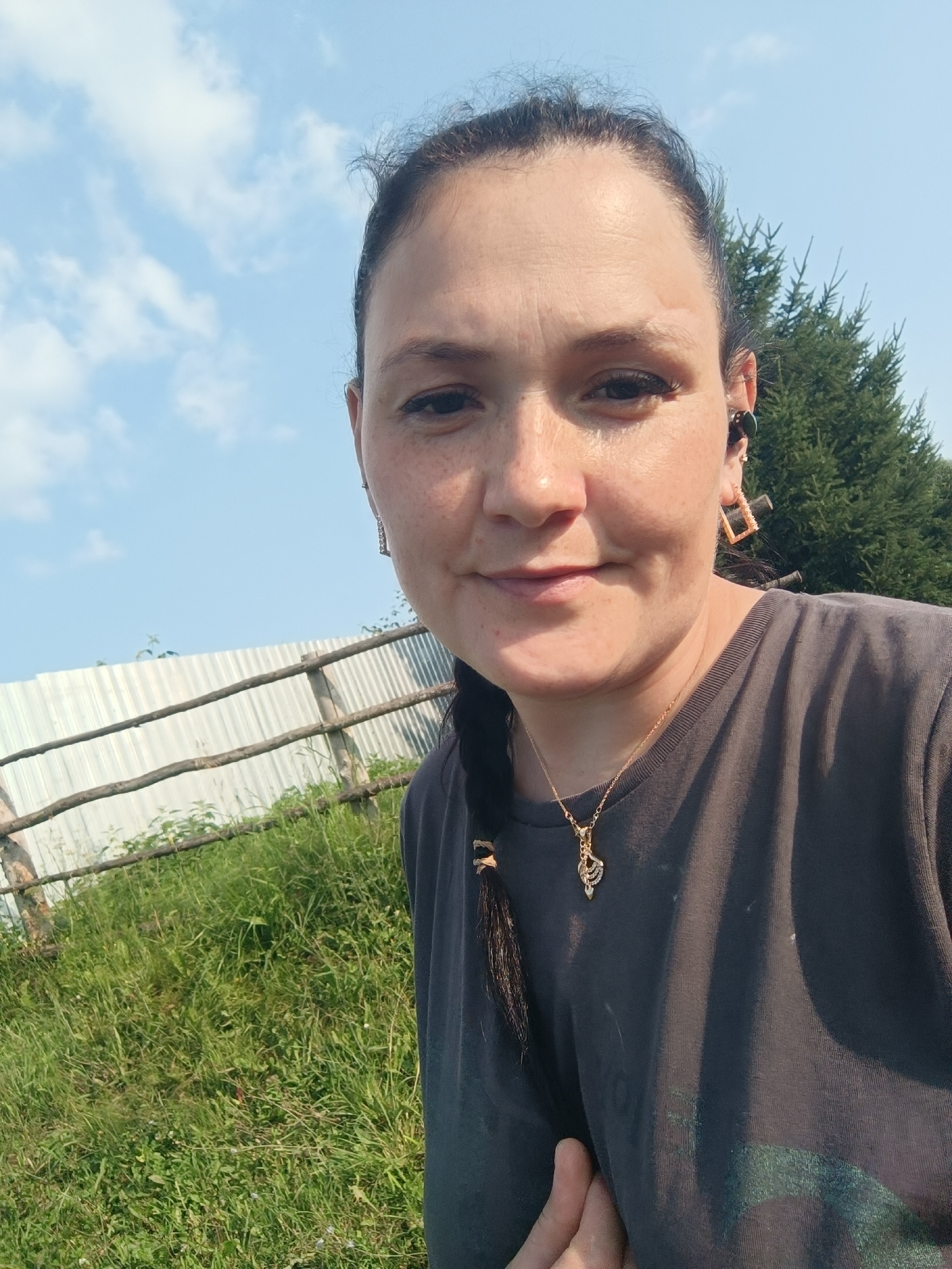 The image is a close-up selfie taken outdoors on a sunny day. The subject is a woman with dark hair pulled back, a braid hanging down her side. She is wearing a dark t-shirt with a gold necklace featuring a pendant. She has on square-shaped earrings and a black earbud. The background features a grassy hill, a rustic wooden fence with corrugated metal sheeting behind it, and evergreen trees. The sky is blue with scattered white clouds. The lighting suggests it is daytime, likely late morning or afternoon. The overall mood appears casual and relaxed. There is no visible text in the image. Based on the location context provided, the scene is Karsha, Russia.