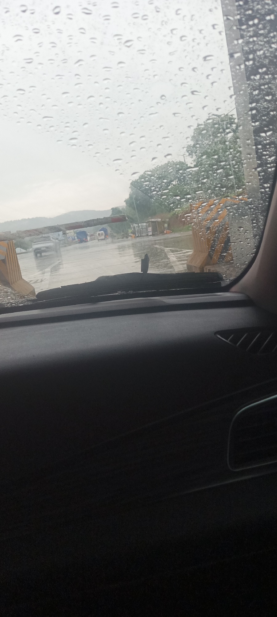 The image shows a view from inside a car, looking out through a rain-streaked windshield. The car is at a road construction area, likely in Kadarko, Nigeria, as noted. There are large yellow construction barriers visible outside. Other vehicles, including trucks and a white van, are visible in the background on the wet road surface. Trees are also visible in the background.