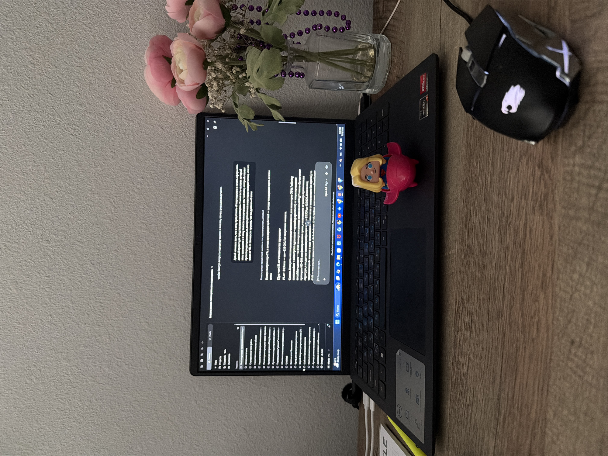 This is a close-up, eye-level, indoor, still photograph of a desk scene. The desk is made of wood grain laminate. On the desk is an open black Dell laptop with a dark screen displaying text in Russian. In front of the laptop is a small, pink, plastic toy airplane with a blonde cartoon character figure. To the right of the laptop is a clear glass vase containing a bouquet of pink peonies and white baby's breath, adorned with a strand of purple beads. In the foreground, to the right of the laptop, is a black computer mouse with silver accents and a white illuminated logo. A yellow highlighter and a white notepad are partially visible on the left side of the desk. The background is a textured, light-colored wall. The lighting appears to be artificial. The image does not provide any information to determine the location in the city of Orlando, United States of America.