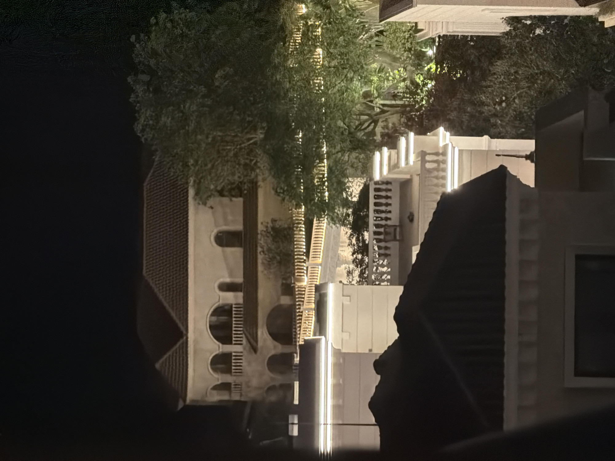This is a night-time photograph showcasing a multi-tiered architectural complex, likely a residential area or resort, located in Unknown, Unknown. The scene is dominated by a large, light-colored building in the background, featuring a dark tiled roof with multiple gables, and several arched windows or loggias across at least two visible stories. These upper levels are adorned with white balustrades.
In the midground, a series of lower structures with flat roofs and intricate balustrades are visible, horizontally illuminated by prominent linear strip lighting. A large, well-lit tree with dense foliage occupies the right side of the midground, partially obscuring the building behind it. Further to the left and foreground, dark rooflines of other buildings are silhouetted against the illuminated structures. Artificial lighting is a prominent feature throughout the scene, highlighting the architectural details, balustrades, and vegetation, creating bright accents against the dark night sky. No individuals are present in the image.